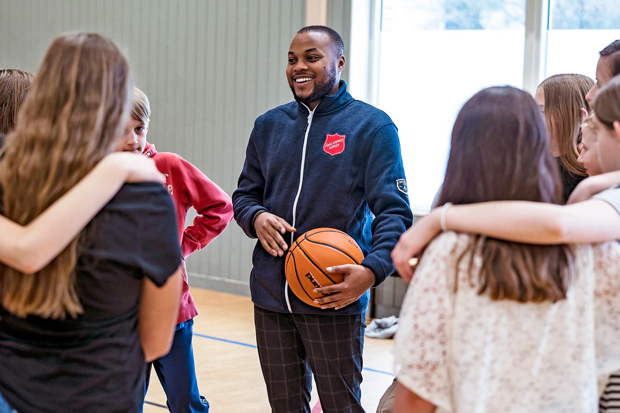 En leende ung man med Frälsningsarméns sköld på tröjan står med en basketboll i händerna i en gymnastiksal. Runt om honom står ungdomar med armarna över varandras axlar.