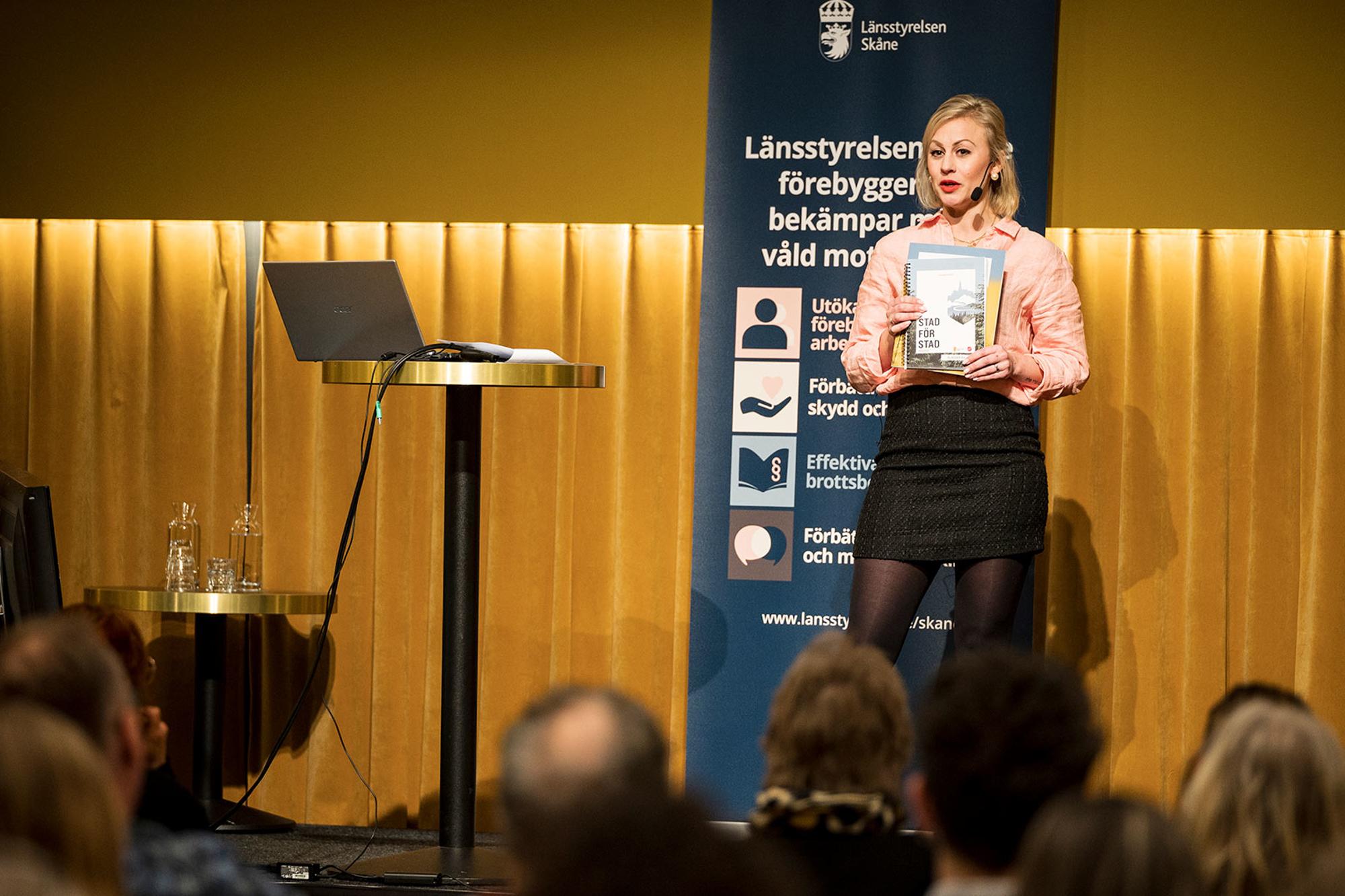 Emma Cotterill står på en scen vid en konferens i Malmö kring människohandel. Hon håller materialet Stad för stad i händerna.