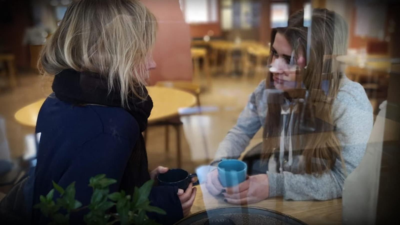 Bild tagen genom ett fönster. På bilden syns två kvinnor som sitter mittemot varandra och samtalar vid ett bord. Kvinnorna håller i varsin kaffemugg.