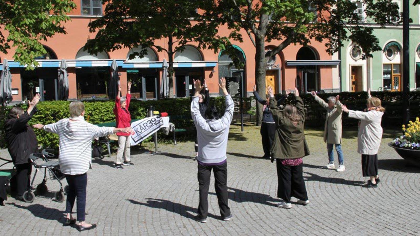 Äldre deltagare på utegympan som arrangeras av Kår 393 på Greta Garbos torg på Södermalm i Stockholm. Deltagarna sträcker armarna upp i luften.