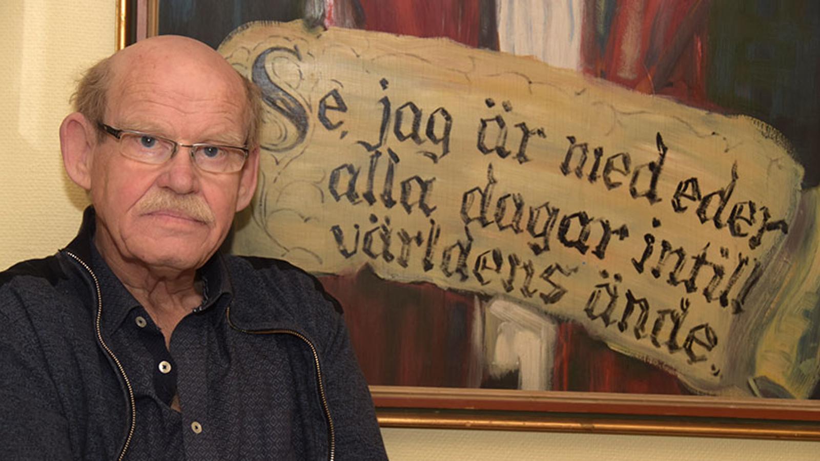 Porträttbild av Karl Gunnar Svensson. Han står till vänster om en tavla med texten "Se, jag är med eder alla dagar intill världens ände", och tittar in i kameran.