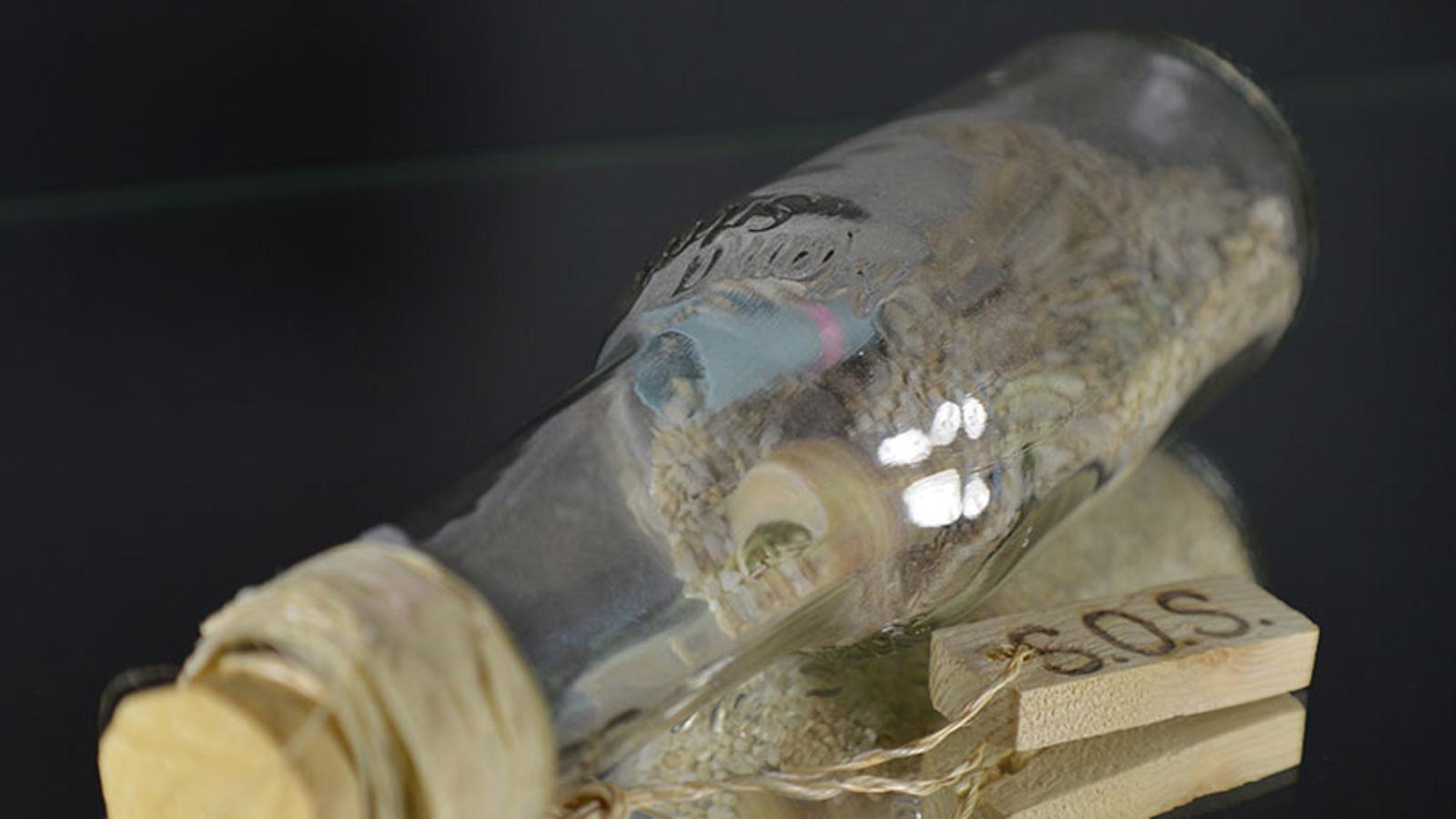 En glasflaska med en lapp i. På flaskan hänger det ett snöre som sitter fast i en liten träbit med texten "SOS".