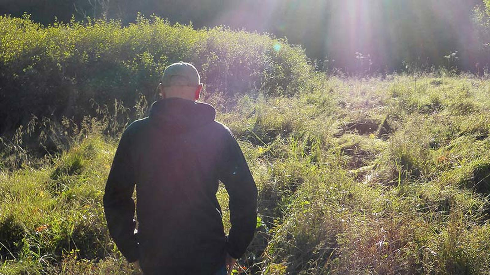 En man med ryggtavlan mot kameran. Han står ute i naturen och i bakgrunden syns solstrålar.