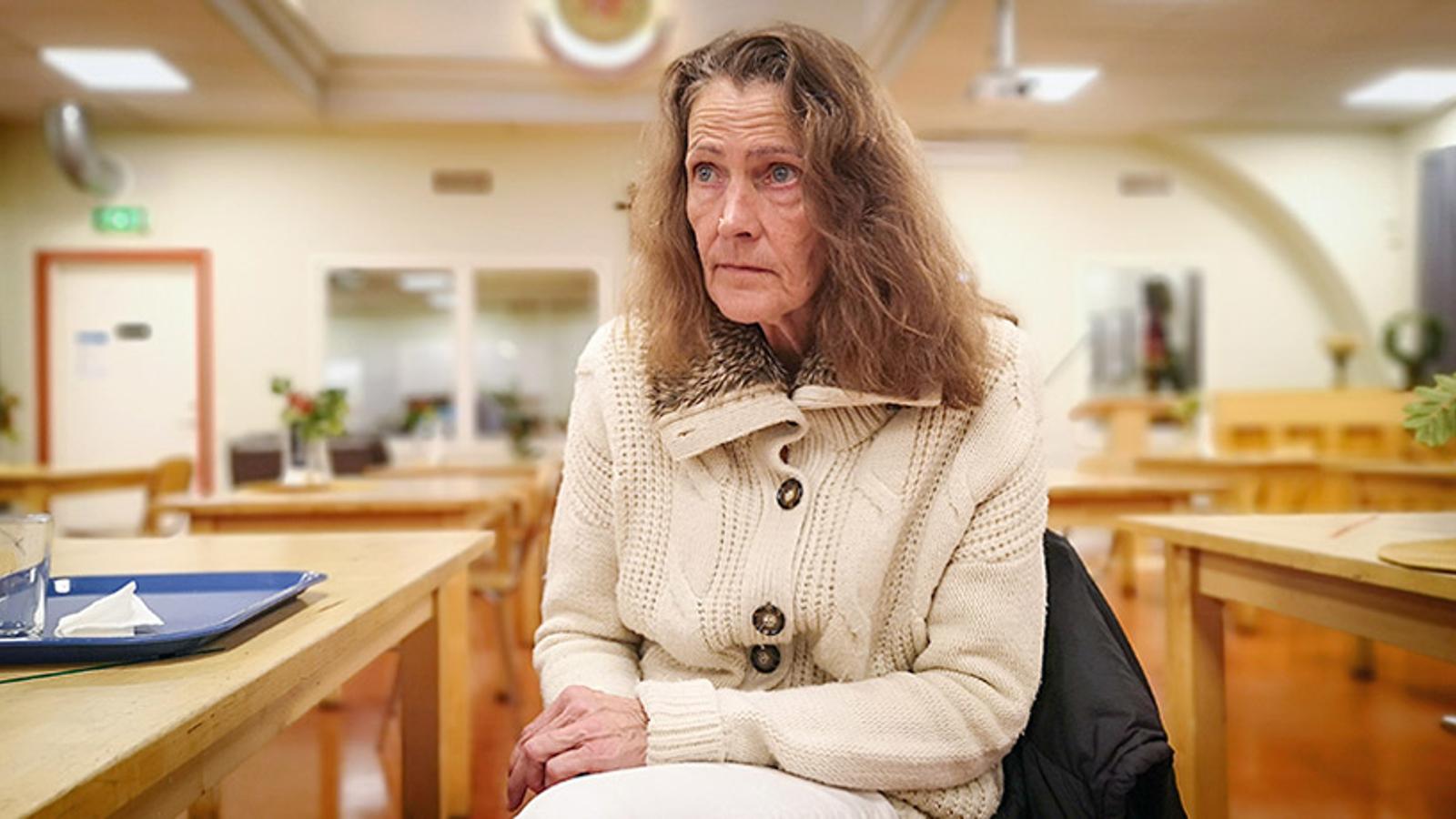 En kvinna, Sussie, som tittar till vänster om kameran. Hon sitter vid ett bord i sociala centrets lokal. På bordet står det en blå bricka och i bakgrunden syns flera bord.