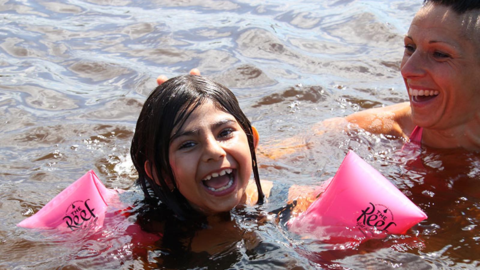 En glad flicka som badar med armpuffar. Bredvid henne badar en kvinna som ler mot flickan.