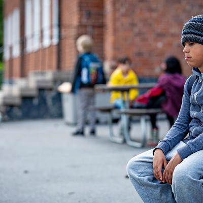 En ung pojke sitter ensam på en bänk på en skolgård. I bakgrunden syns fyra andra barn som leker.