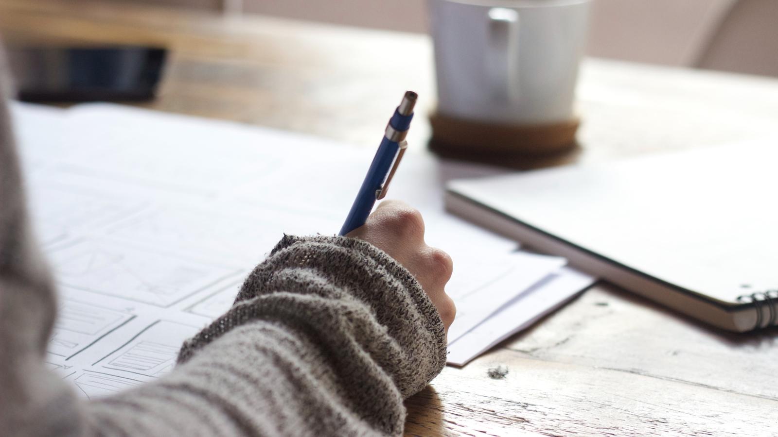 En hand i fokus som håller i en penna. Handen ligger på ett bord med papper och ett block på.