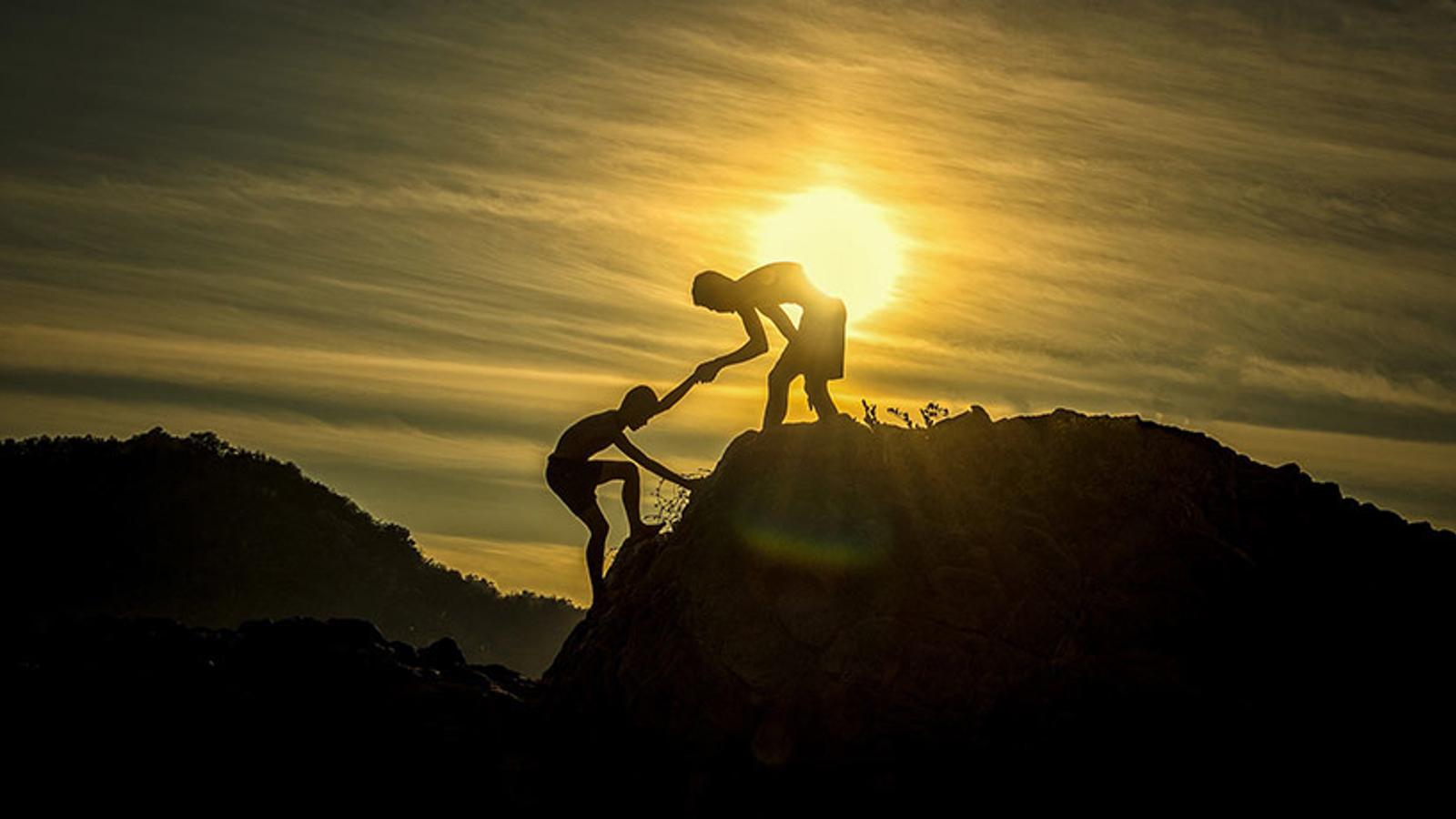 En person som får hjälp upp för ett berg av en annan person, och med en solnedgång i bakgrunden.