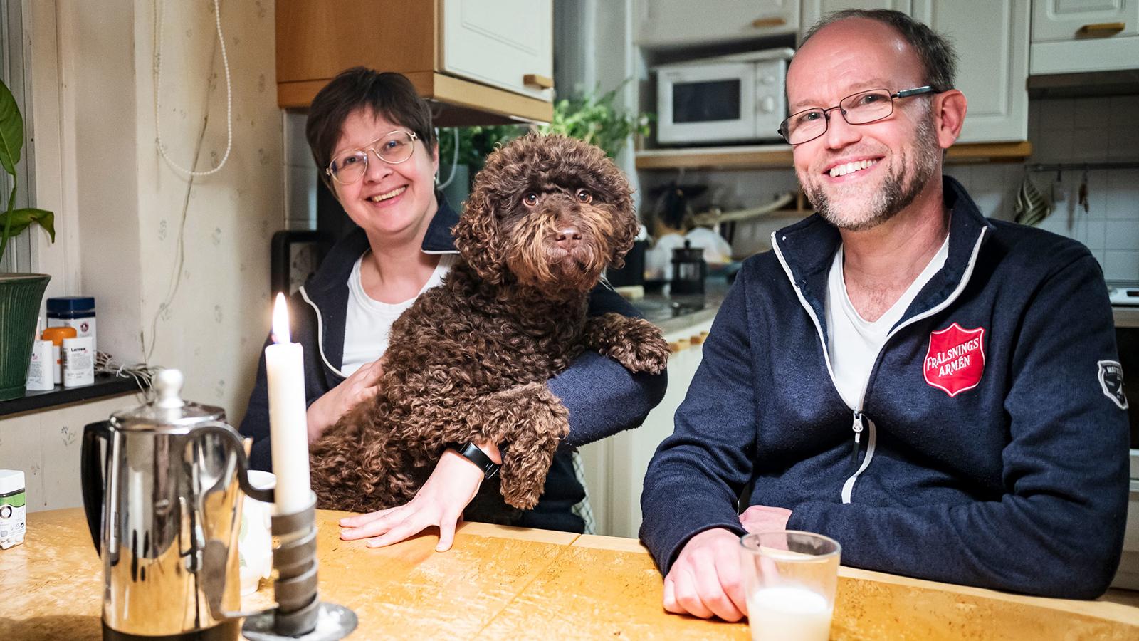 Carolina och Andreas hemma vid köksbordet tillsammans med familjens hund. Bakom dem syns ett kök.