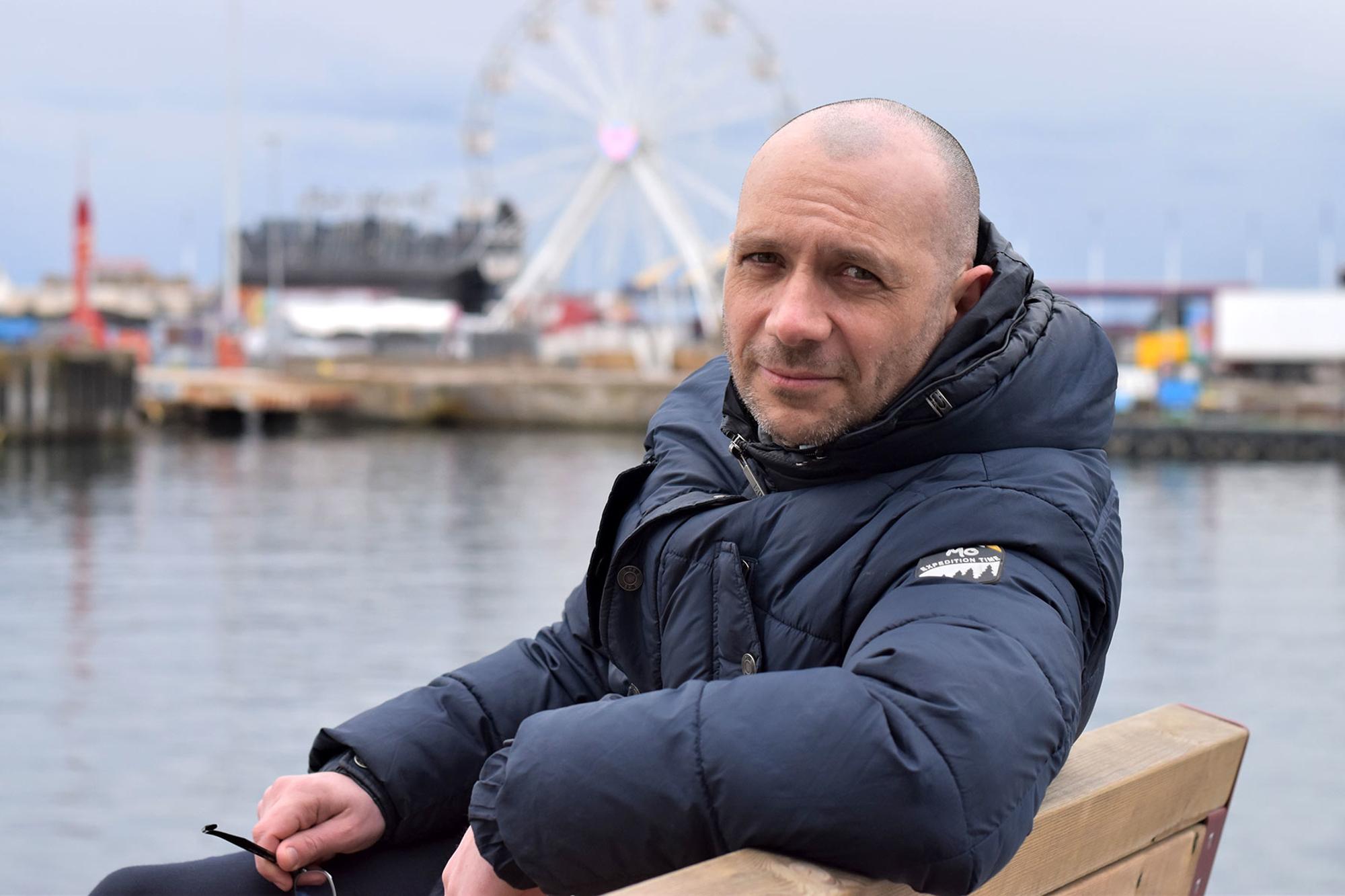Porträttbild av Mark Cotterill. Han sitter på en bänk med blicken i kameran, och bakom honom syns en hamn.
