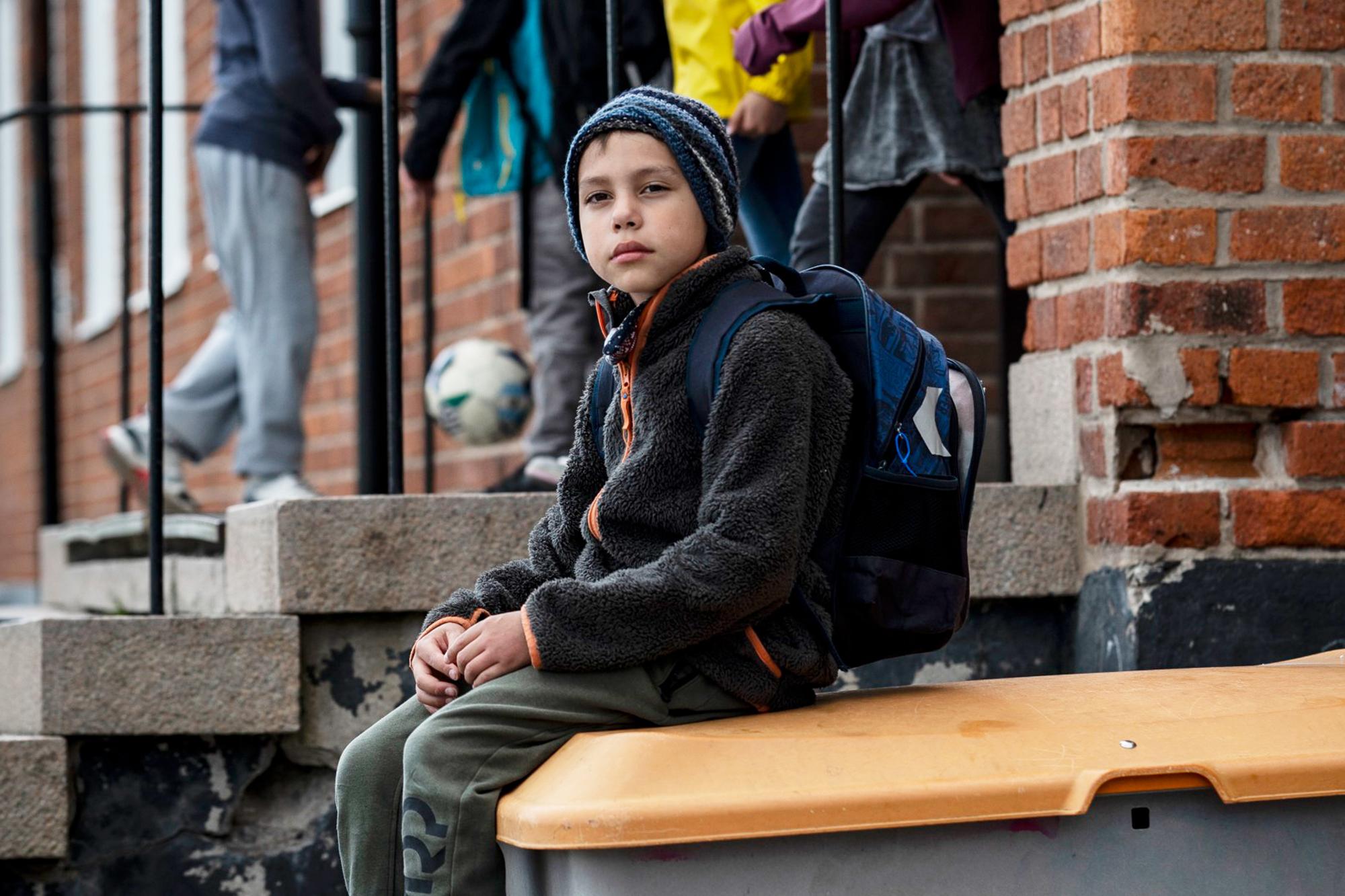 En pojke med ryggsäck sitter på en låda på en skolgård och ser uppgiven ut. Man ser andra barn i bakgrunden.