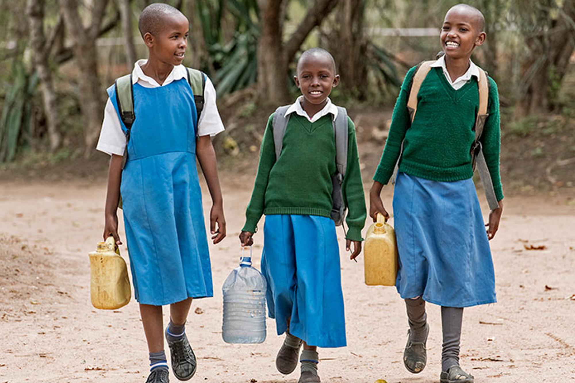 Tre glada flickor i Kenya går tillsammans med ryggsäck på ryggen med varsin plastdunk i handen som de ska fylla med vatten.