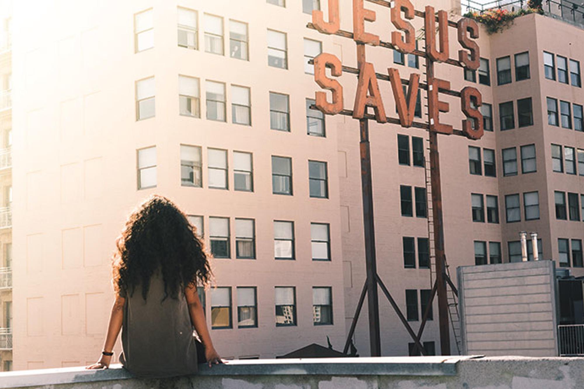 Ryggtavlan på en kvinna som sitter på kanten av en stenmur med blicken mot en byggnad med texten "Jesus Saves". 