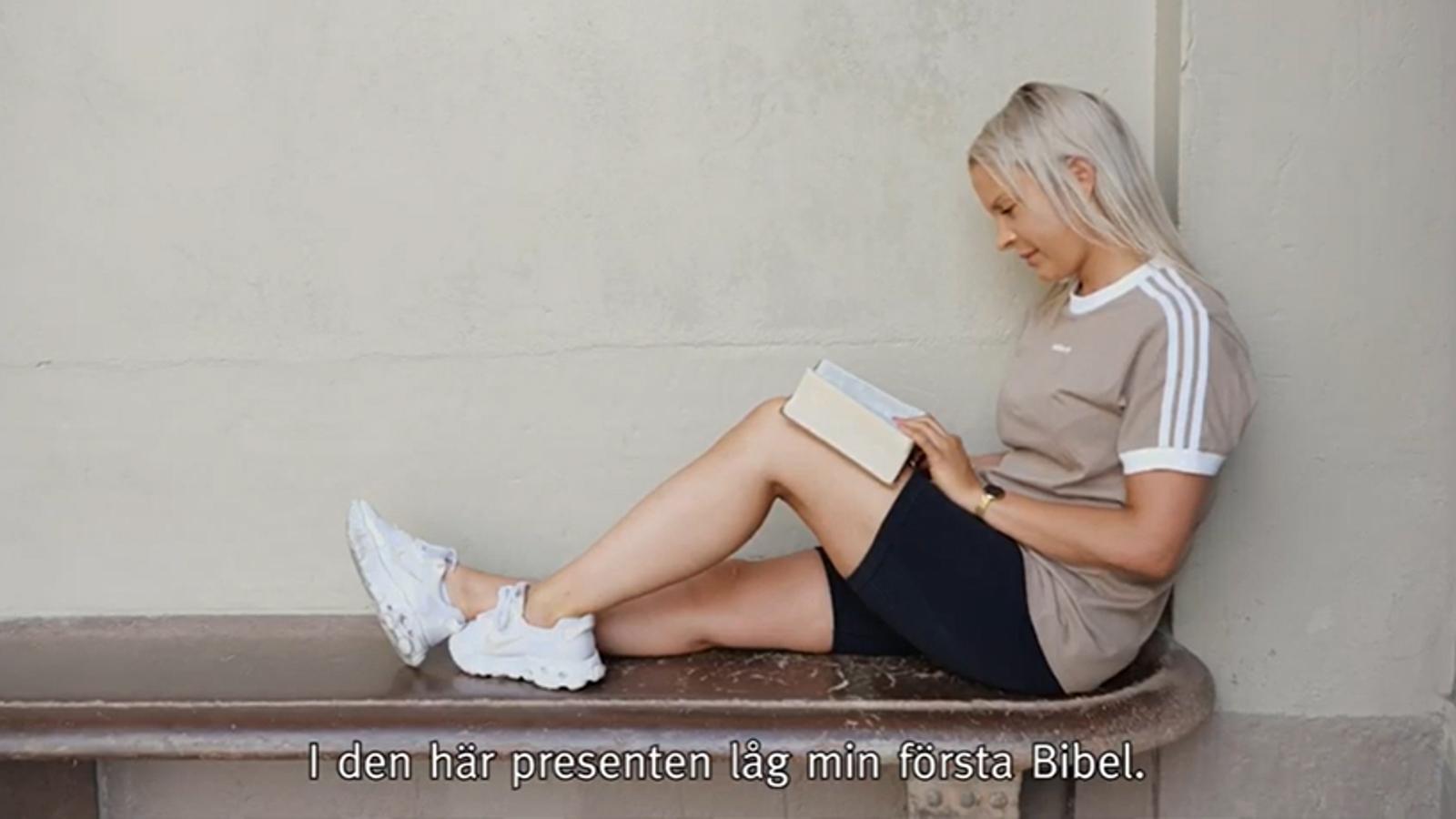 Carolina sitter på en bänk med ryggen lutad mot en vägg. I knät har hon en uppslagen bibel som hon läser i.