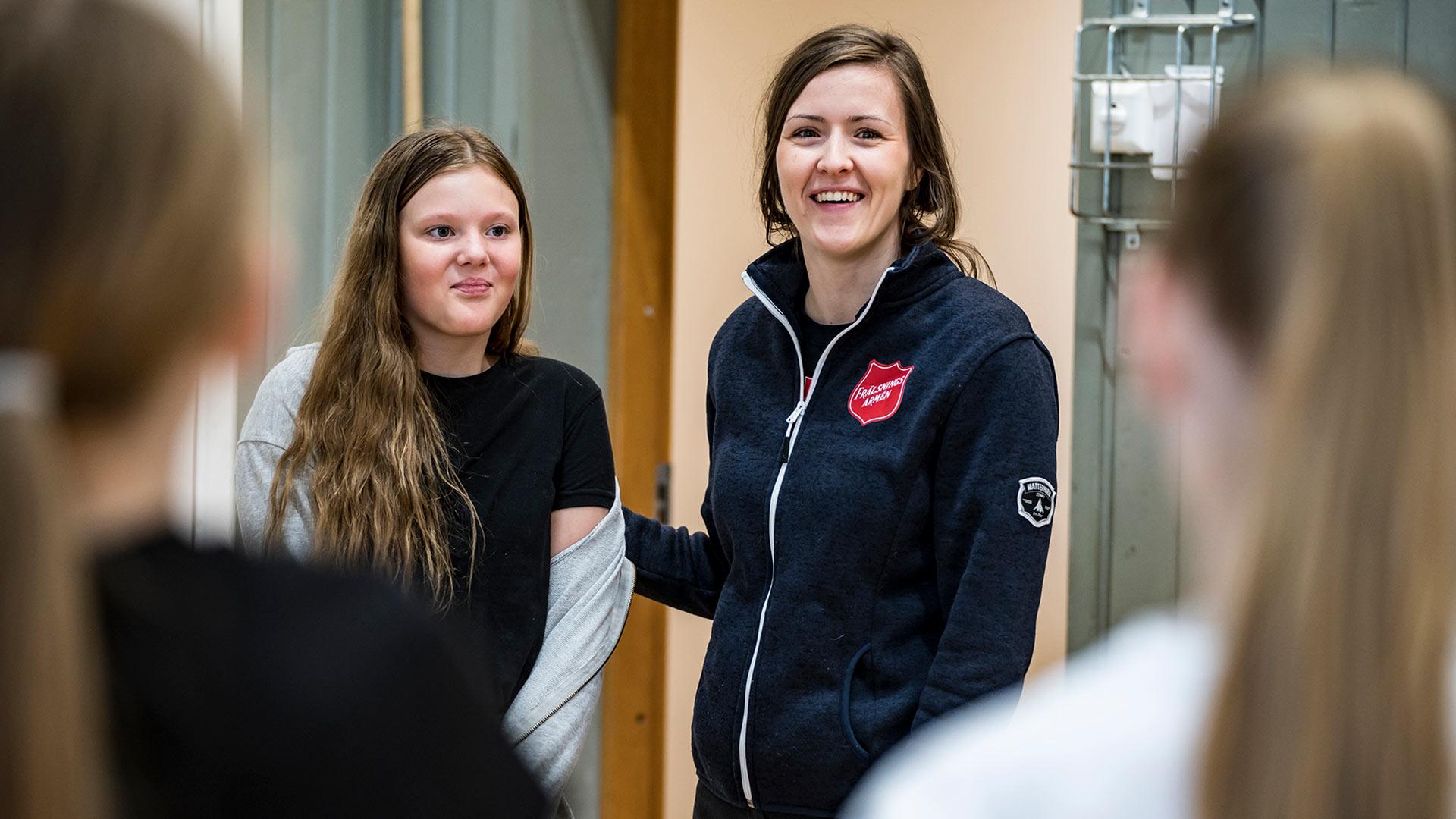 En ung kvinna med Frälsningsarméns sköld på tröjan står tillsammans med en flicka i en dörröppning - de ler emot ett par andra flickor som vi ser ryggtavlorna på.