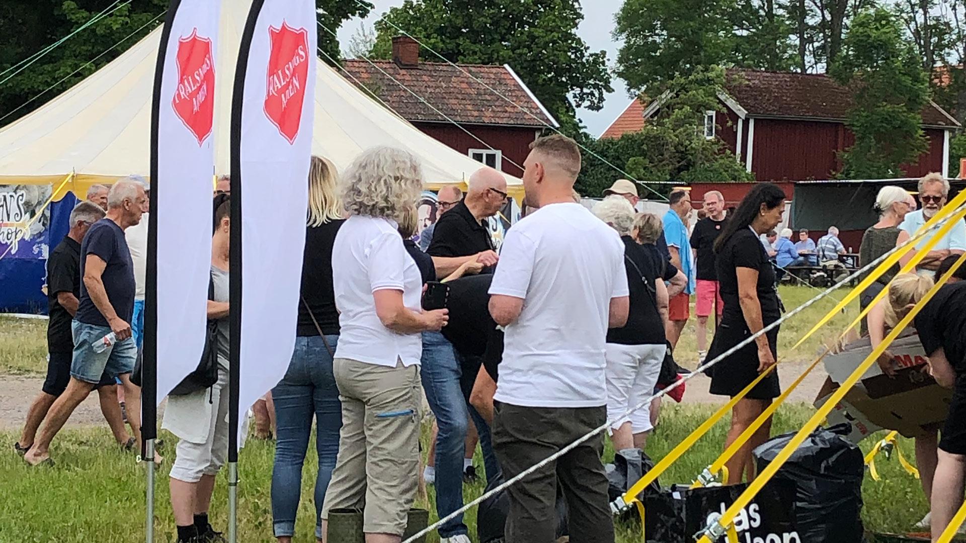 En grupp människor samtalar utomhus invid beachflagga med Frälsningsarméns sköld. Människor i bakgrunden och ett stort tält.