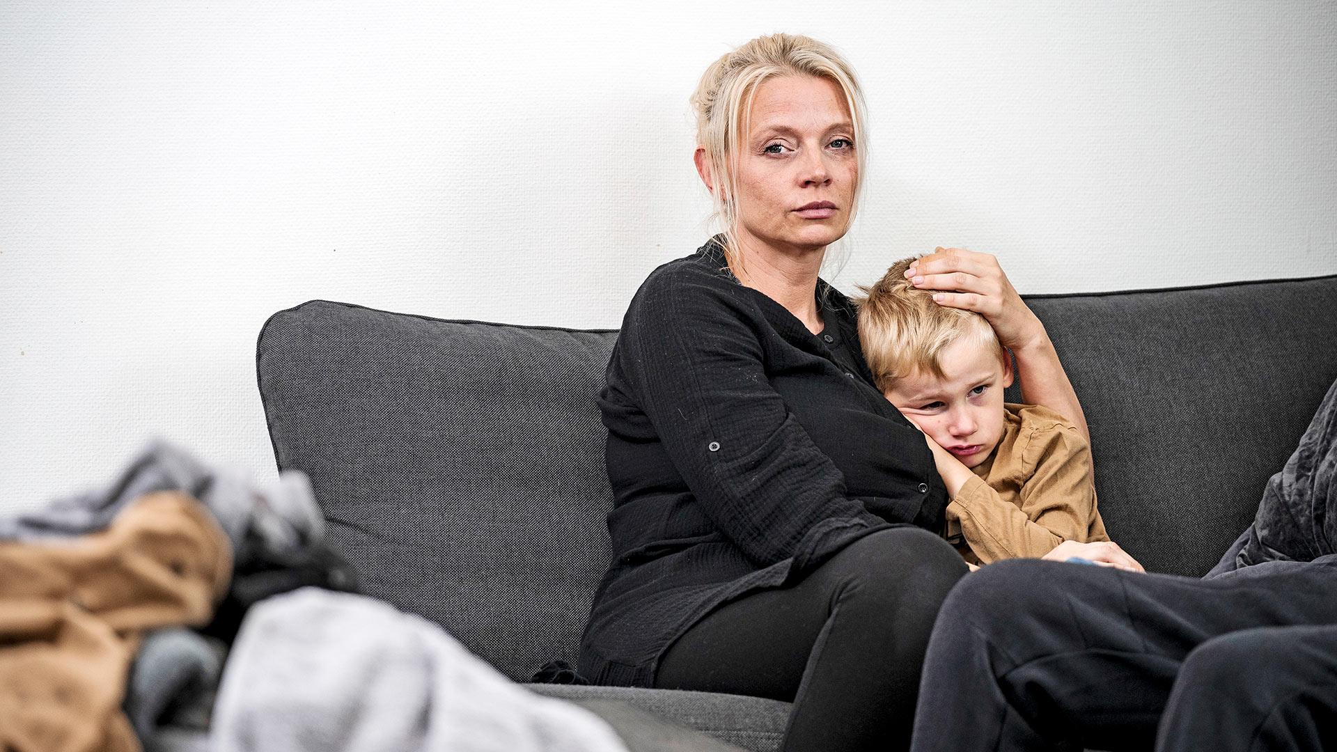 En mamma sitter i en soffa med en liten pojke bredvid sig som hon håller om. Bägge ser allvarliga och ledsna ut.