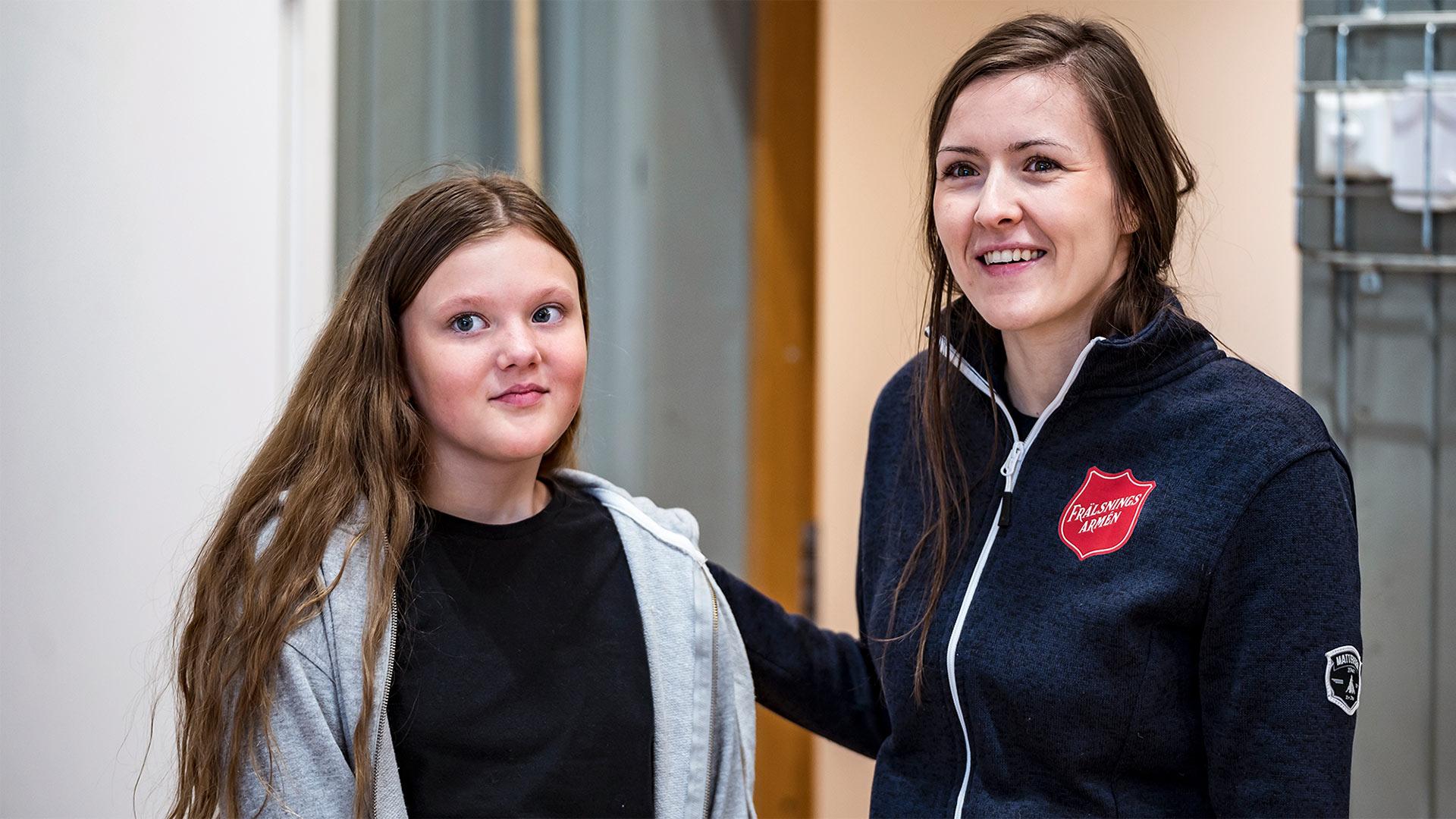 En ung kvinna med Frälsningsarméns sköld på tröjan står tillsammans med en flicka i en dörröppning - båda två ser glada ut.