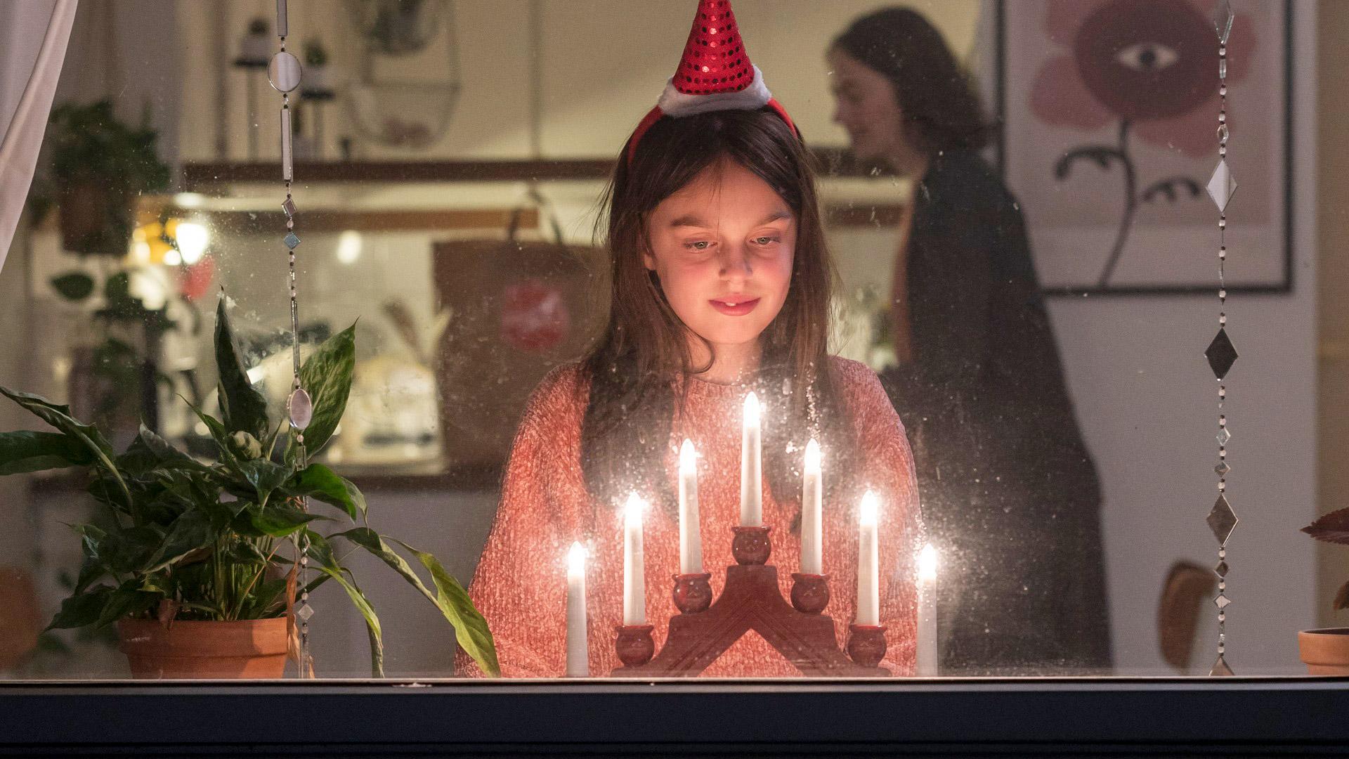En flicka står i ett fönster vid en lysande adventsljusstake. I bakgrunden syns en mamma i köket med en matkasse med Frälsningsarméns sköld på
