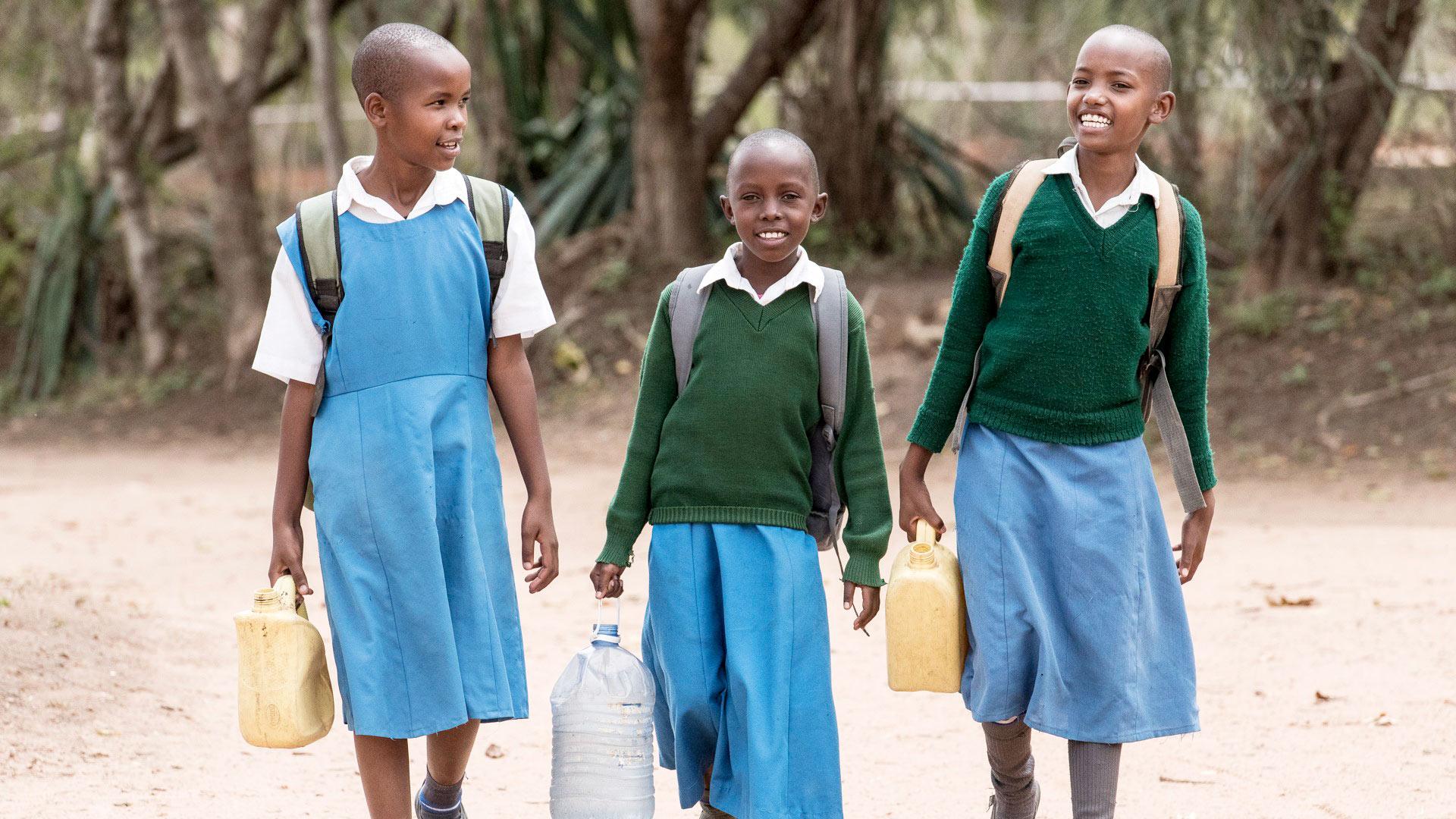 Tre glada flickor i Kenya går tillsammans med ryggsäck på ryggen med varsin plastdunk i handen som de ska fylla med vatten.
