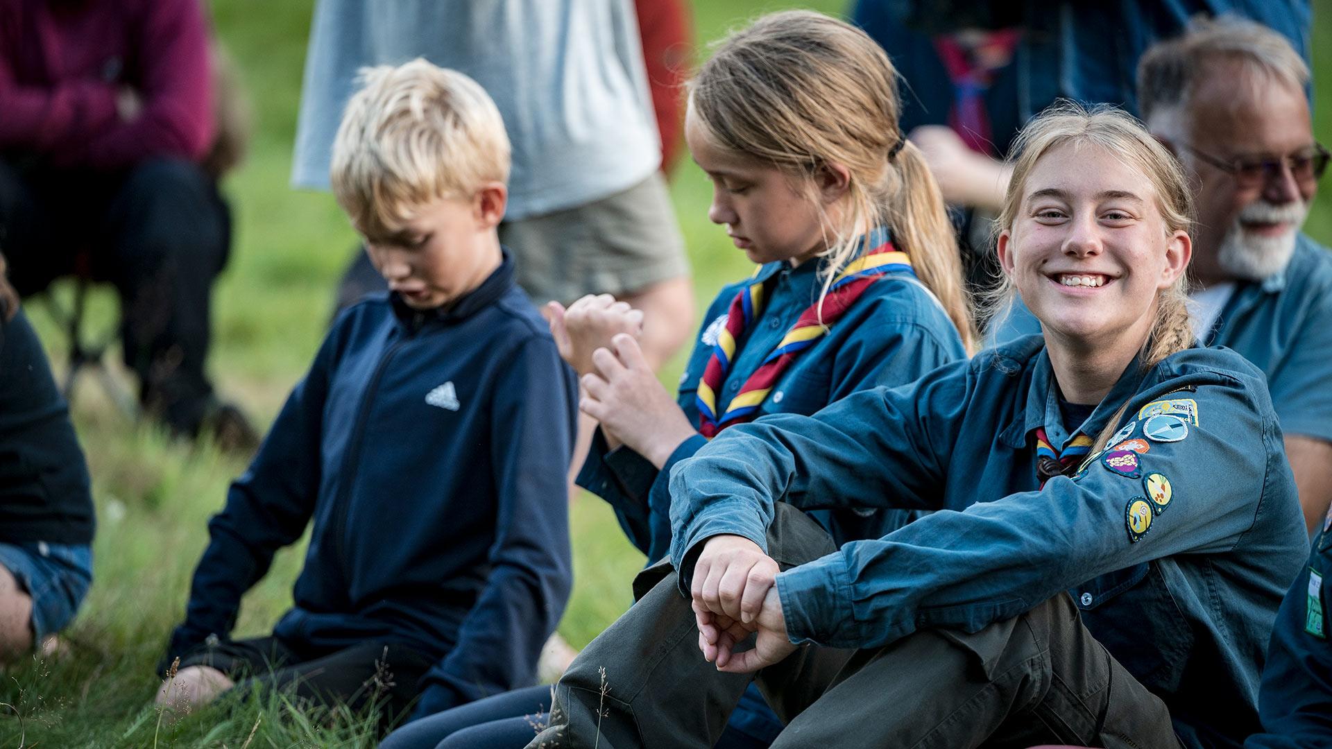 Barn och ungdomar sitter på en gräsmatta, några har scoutkläder på sig.