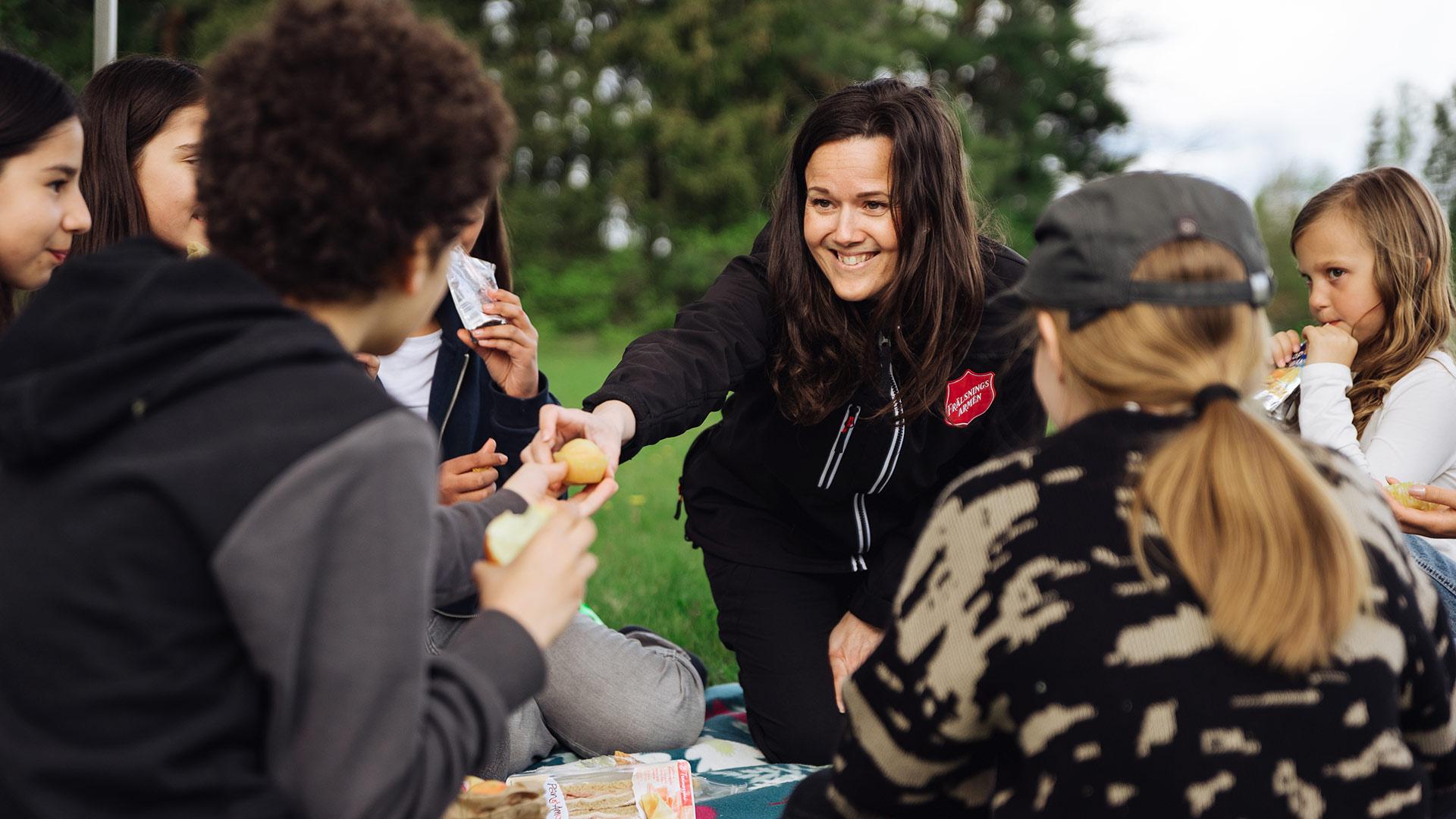 Några ungdomar som sitter i en ring på en gräsmatta - skog syns i bakgrunden. En kvinna med Frälsningsarméns sköld på jackan delar ut fika till ungdomarna.
