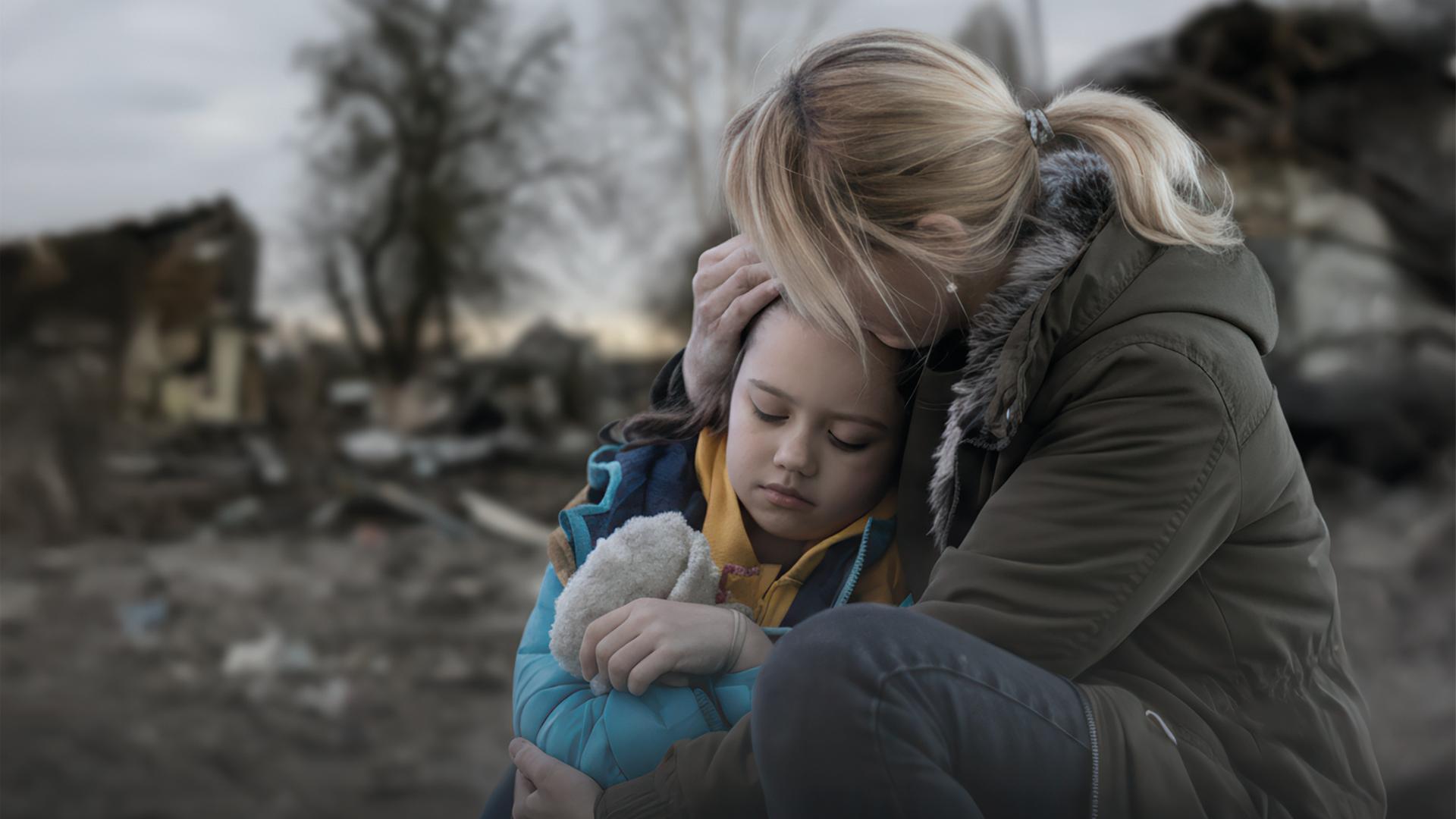 En kvinna i Ukraina håller om sin dotter, i bakgrunden syns flera sönderbombade hus.