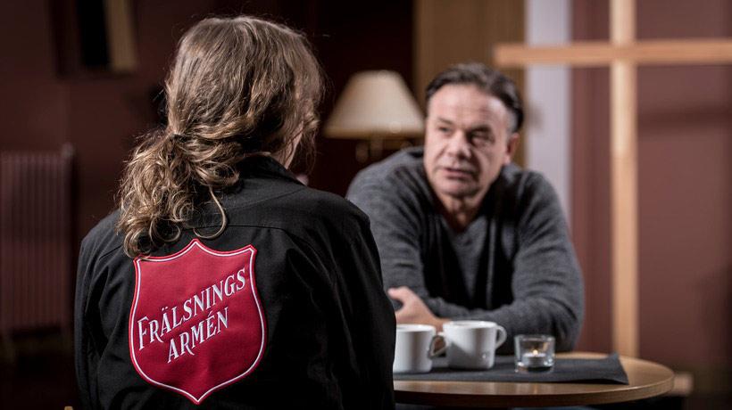 En kvinna från Frälsningsarmén med ryggtavlan mot kameran. Kvinnan sitter vid ett bord och samtalar med en man som sitter mittemot. På bordet står det två kaffemuggar och ett ljus.
