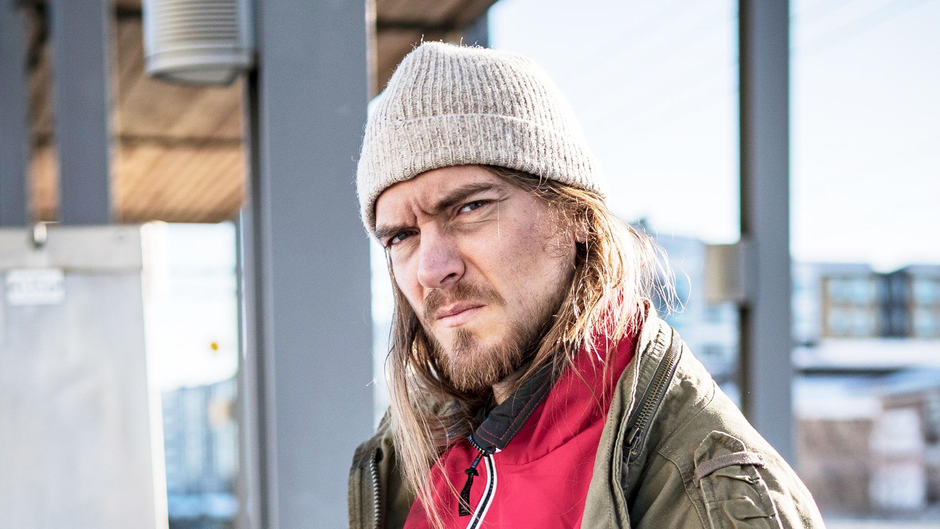 En man med långt hår och skägg sitter på en tågperrong i vinterkläder.