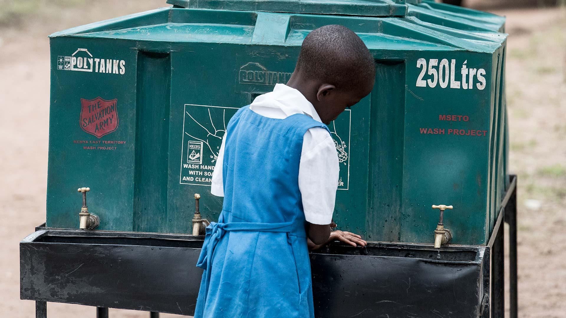 En flicka i skoluniform i Kenya står och tvättar händerna vid en vattentank.