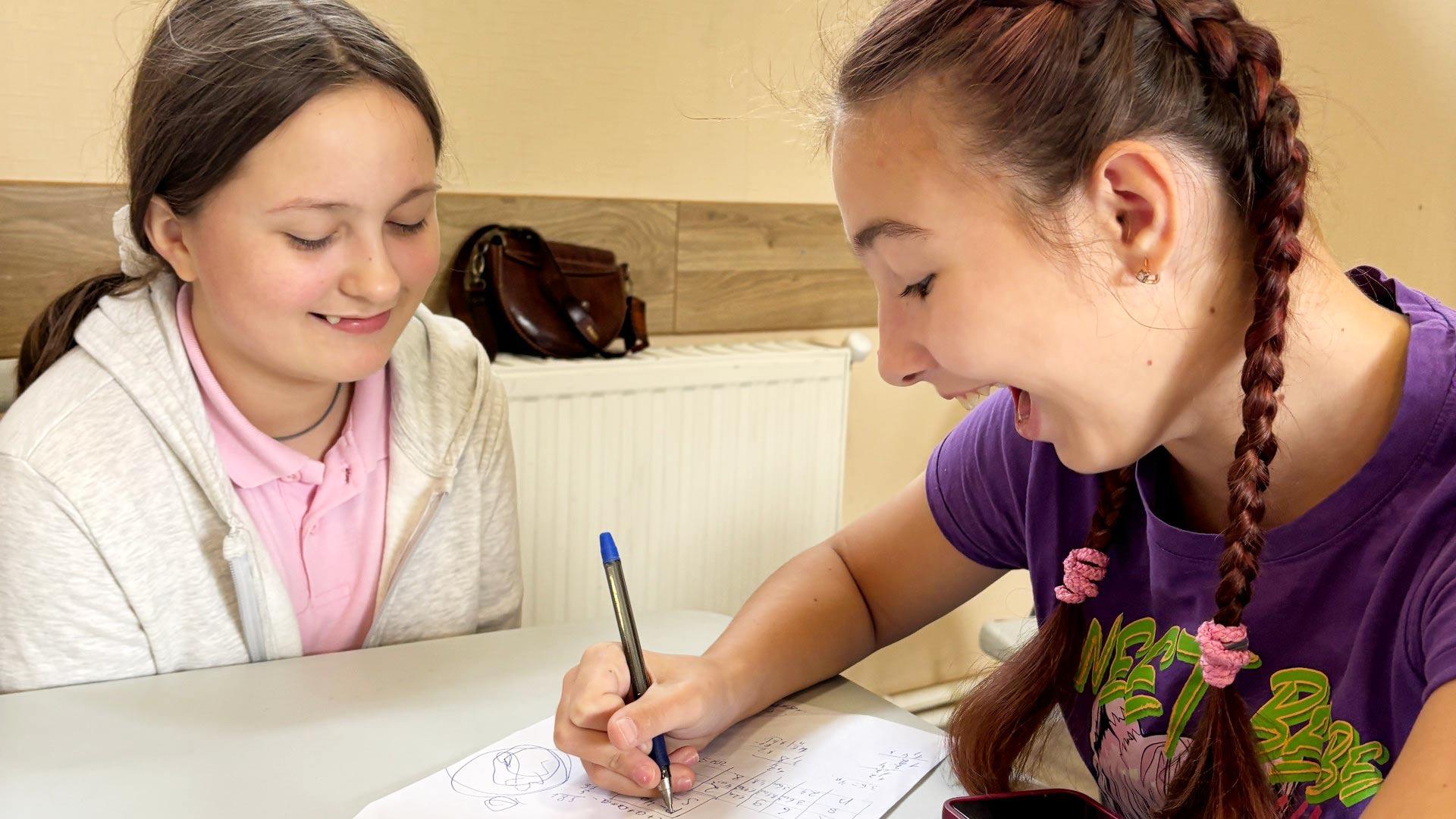 En flicka sitter och skriver på ett papper - en annan flicka sitter mitt emot. Bägge ser glada ut och ler/skrattar.