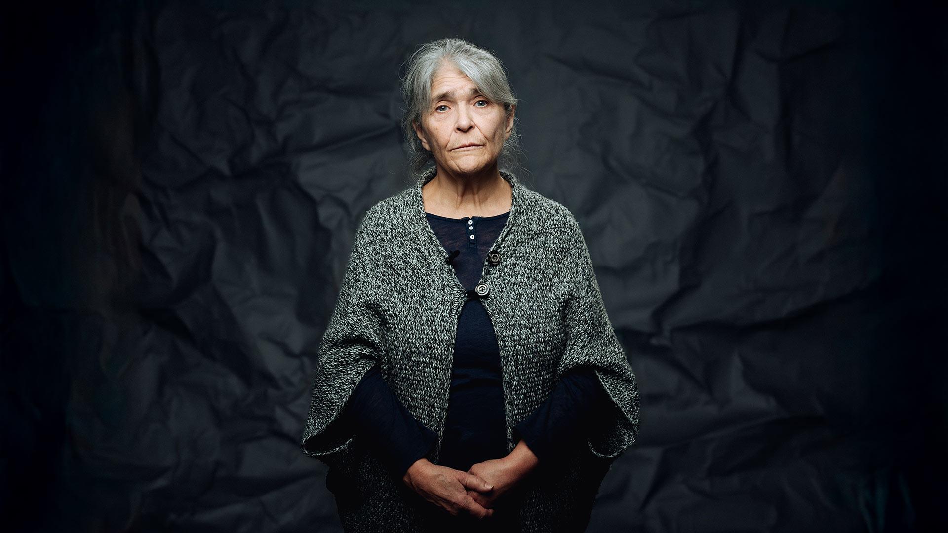 En äldre kvinna med blicken in i kameran - hon ser allvarlig och lite ledsen ut.