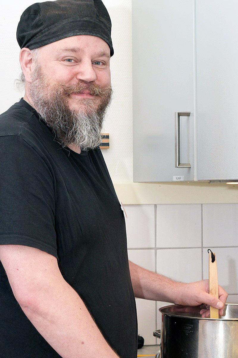 En glad Mattias Nordin iklädd kockkläder som står i ett kök och rör i en gryta med blicken in i kameran.