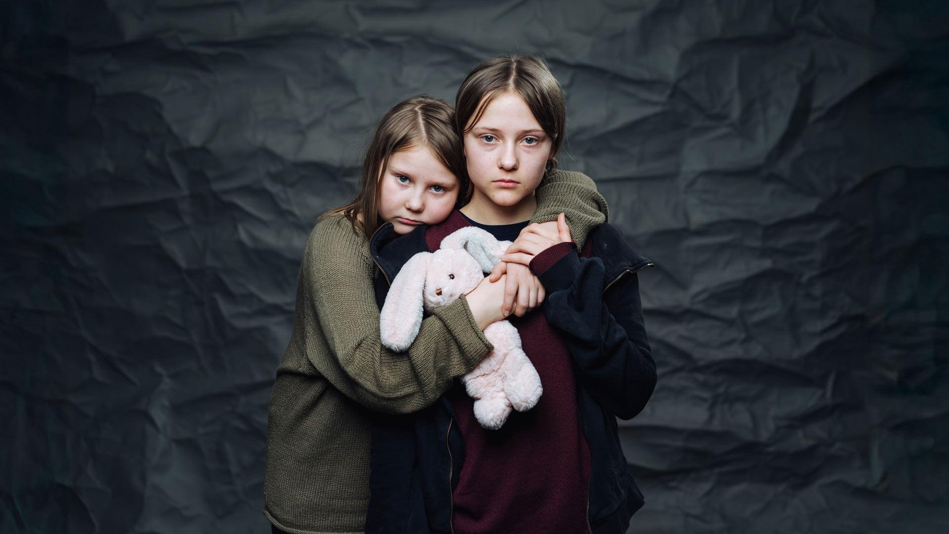 Två allvarliga flickor håller om varandra - i famnen har de ett mjukisdjur. Bakgrunden är svart.