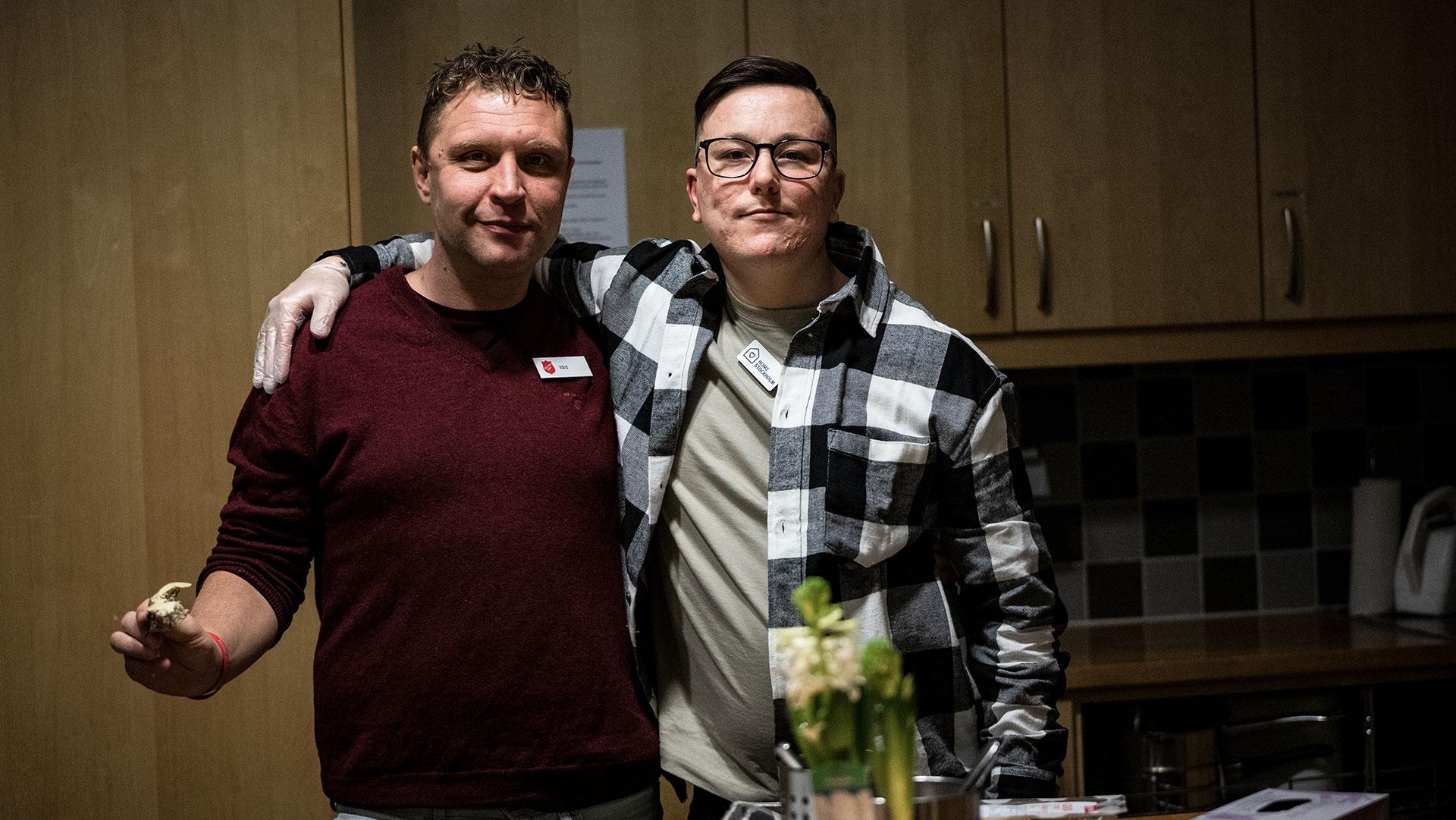 Två unga män står i ett kök med armarna över varandras axlar och tittar in i kameran.