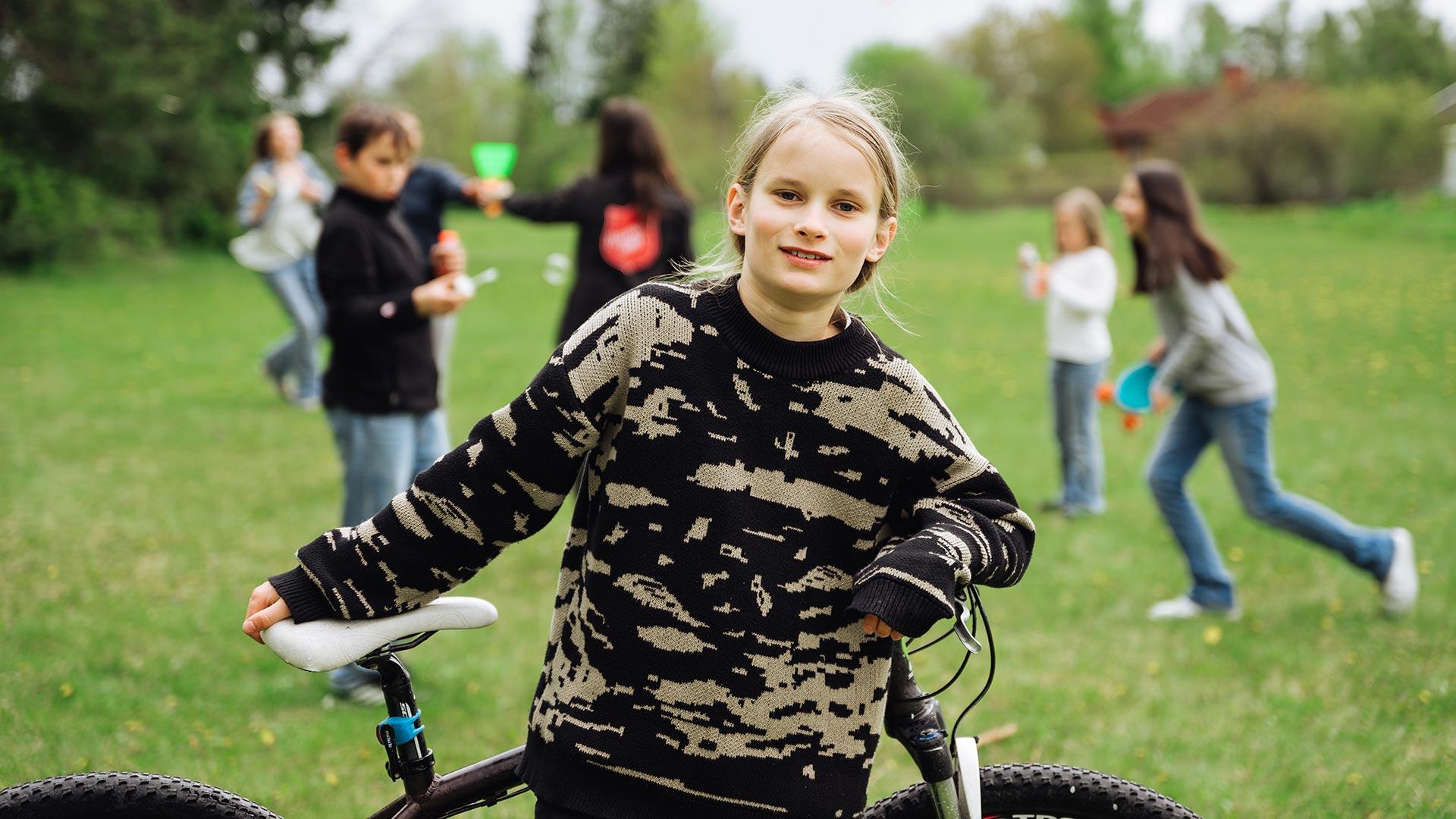 En ung flicka i förgrunden som håller i en cykel. I bakgrunden syns flera ungdomar på en gräsmatta som gör olika aktiviteter.