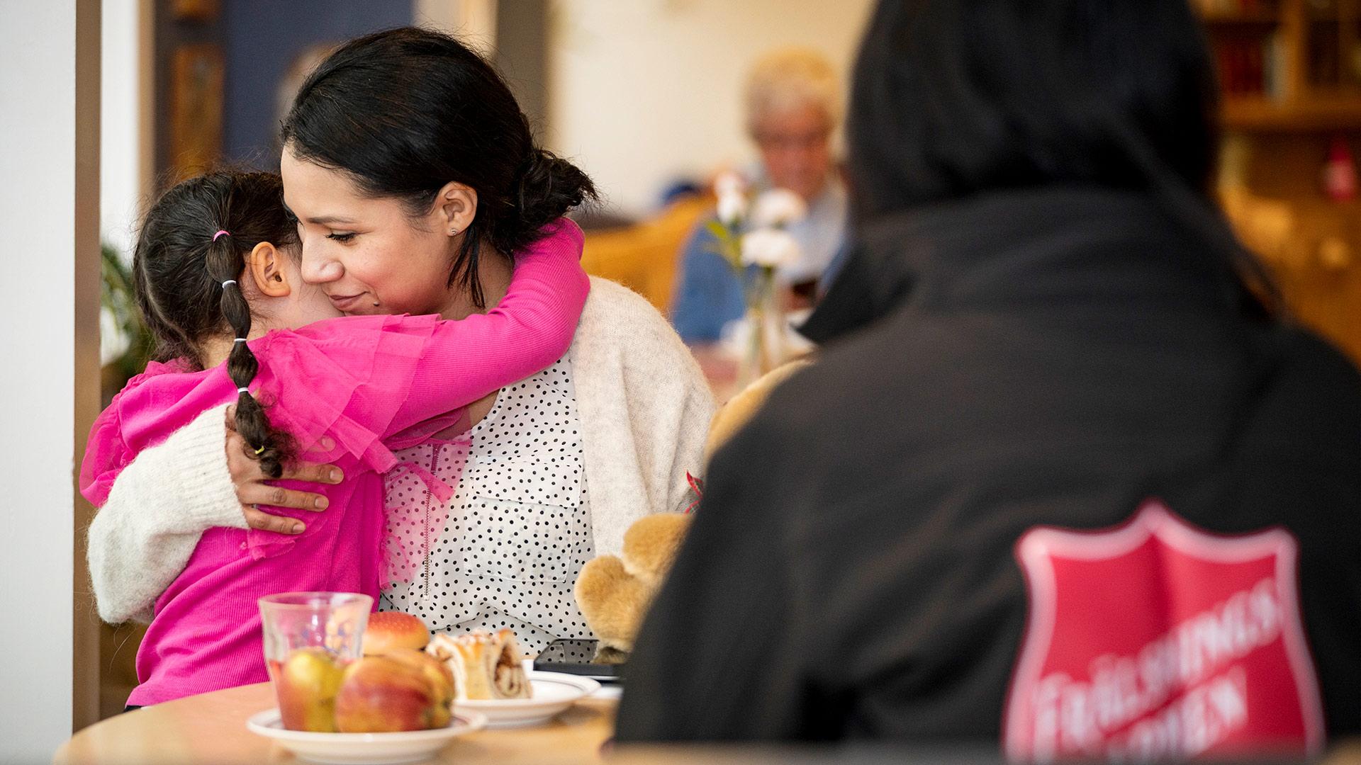 En kvinna kramar om en liten flicka i sitt knä vid ett fikabord med människor i bakgrunden. I förgrunden syns en rygg med Frälsningsarméns sköld.