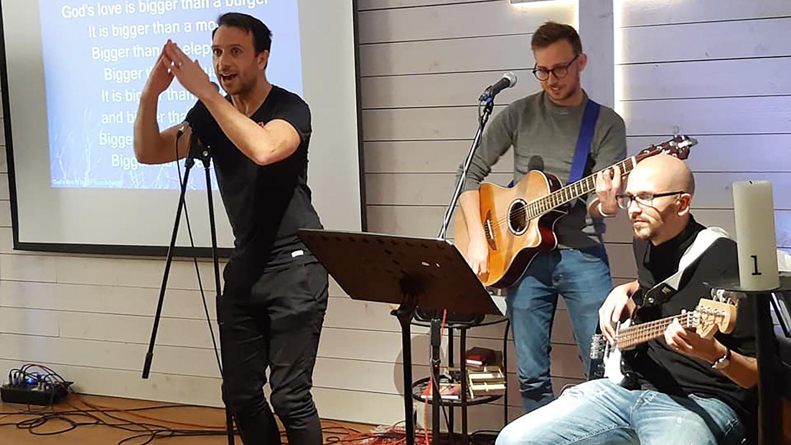 Sång och dans på kåren i Helsingborg. Mark står vid en mikrofon och sjunger, medan två män spelar gitarr och bas.