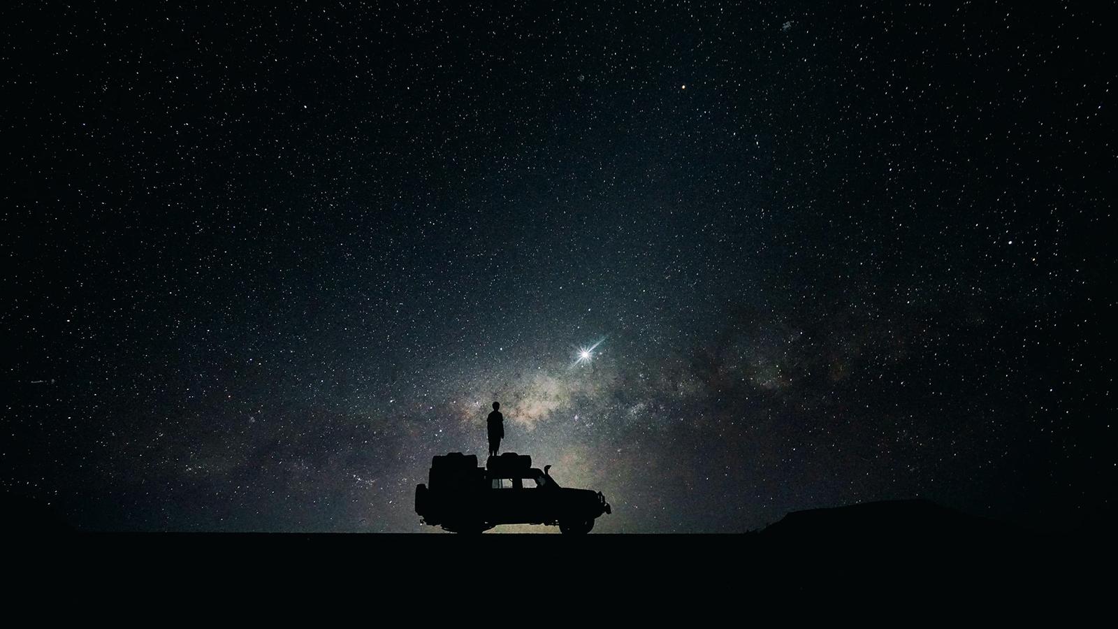 En man står på ett biltak under en stjärnhimmel.