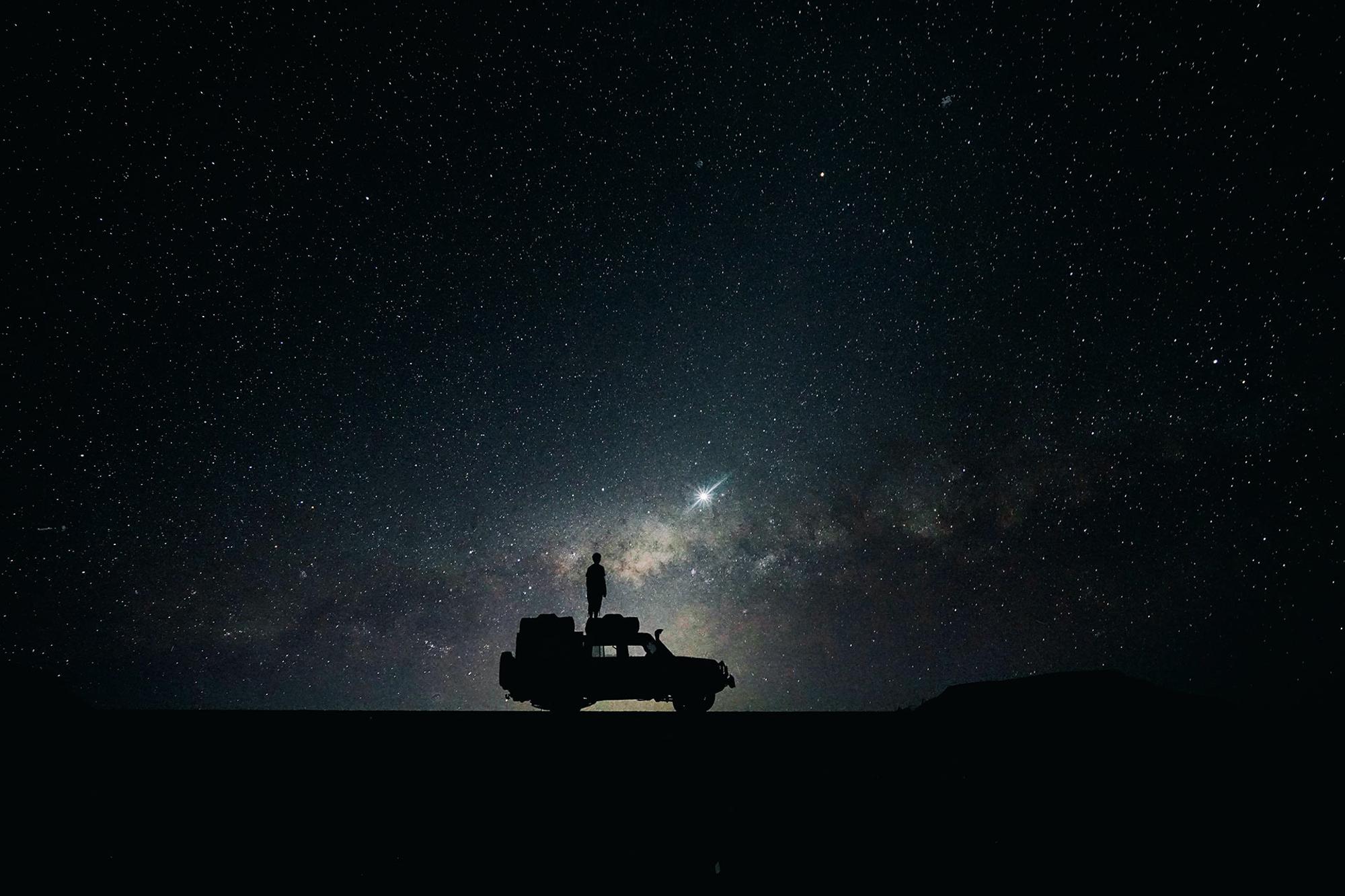 En man står på ett biltak under en stjärnhimmel.