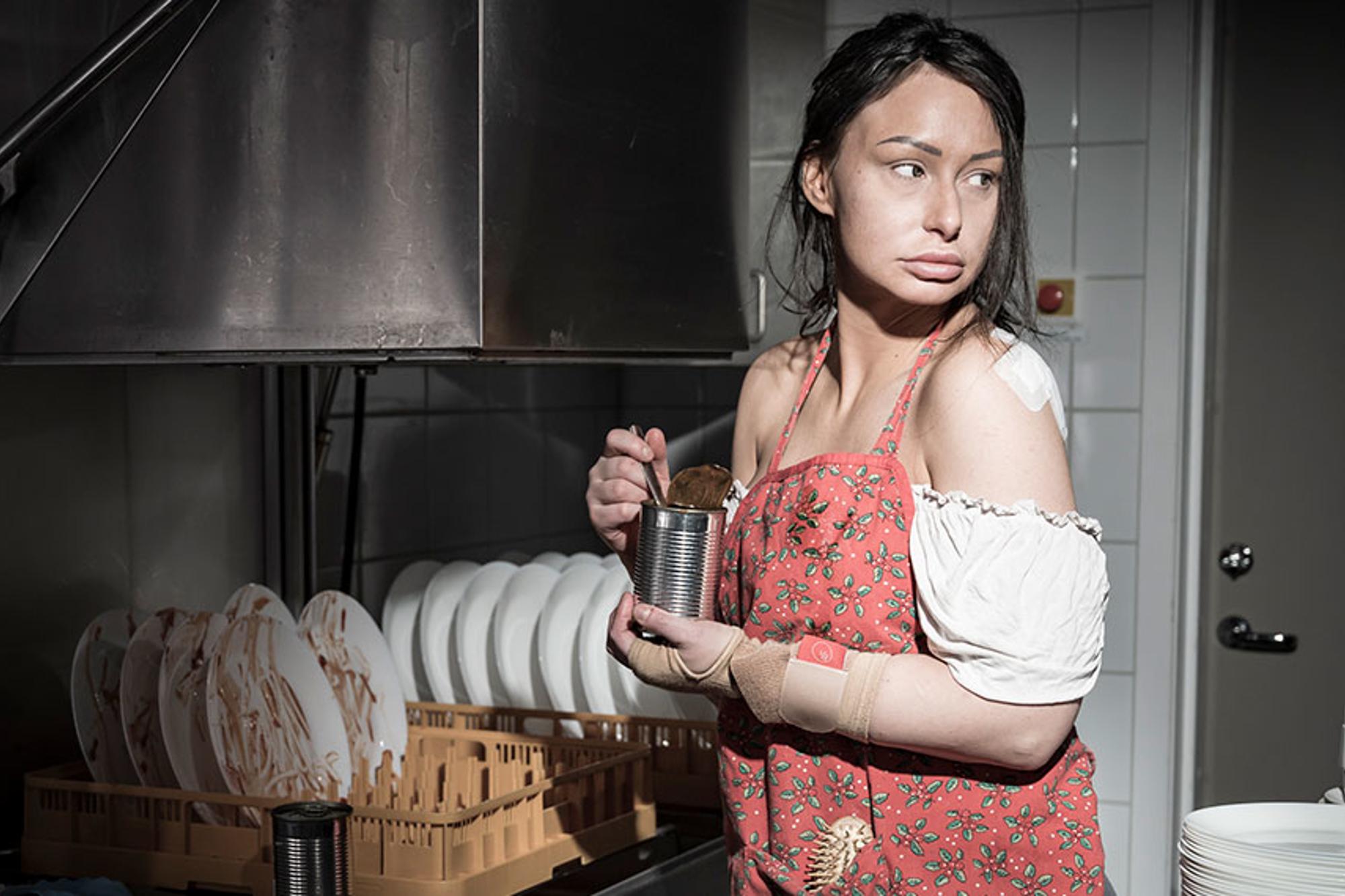 En ung kvinna med plåster på axeln och trasiga kläder står vid en industridiskmaskin och äter ur en konsvervburk.