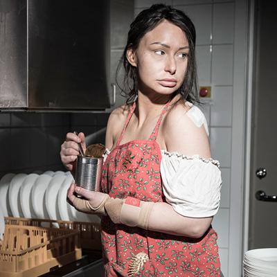 En ung kvinna med plåster på axeln och trasiga kläder står vid en industridiskmaskin och äter ur en konsvervburk.