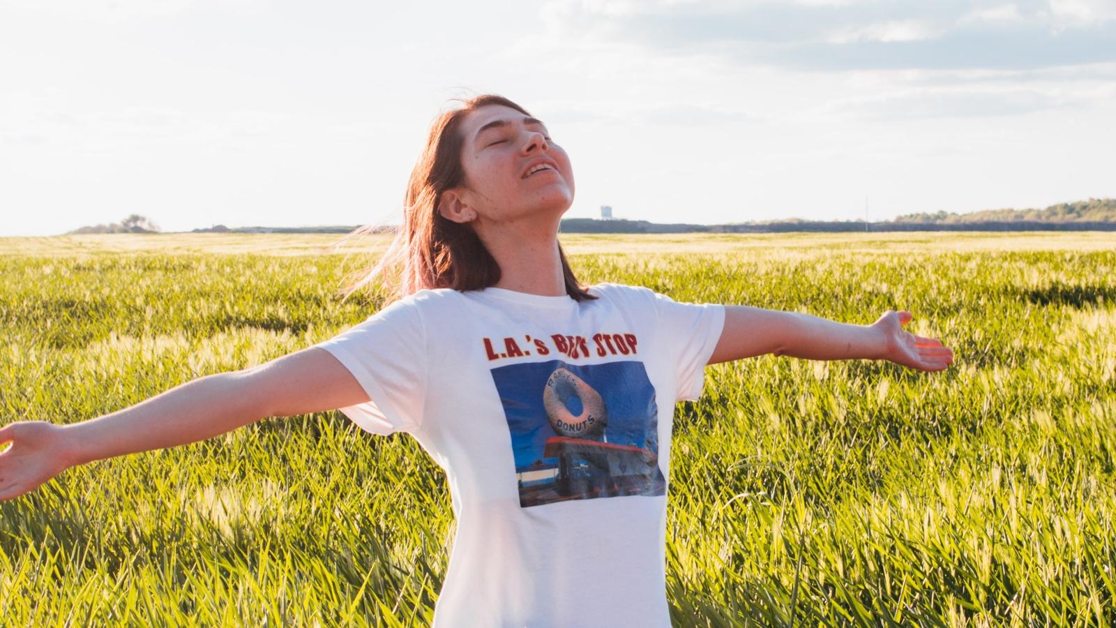 En kvinna som står på en äng av högt gräs och sträcker ut armarna i luften och huvudet upp mot himlen.