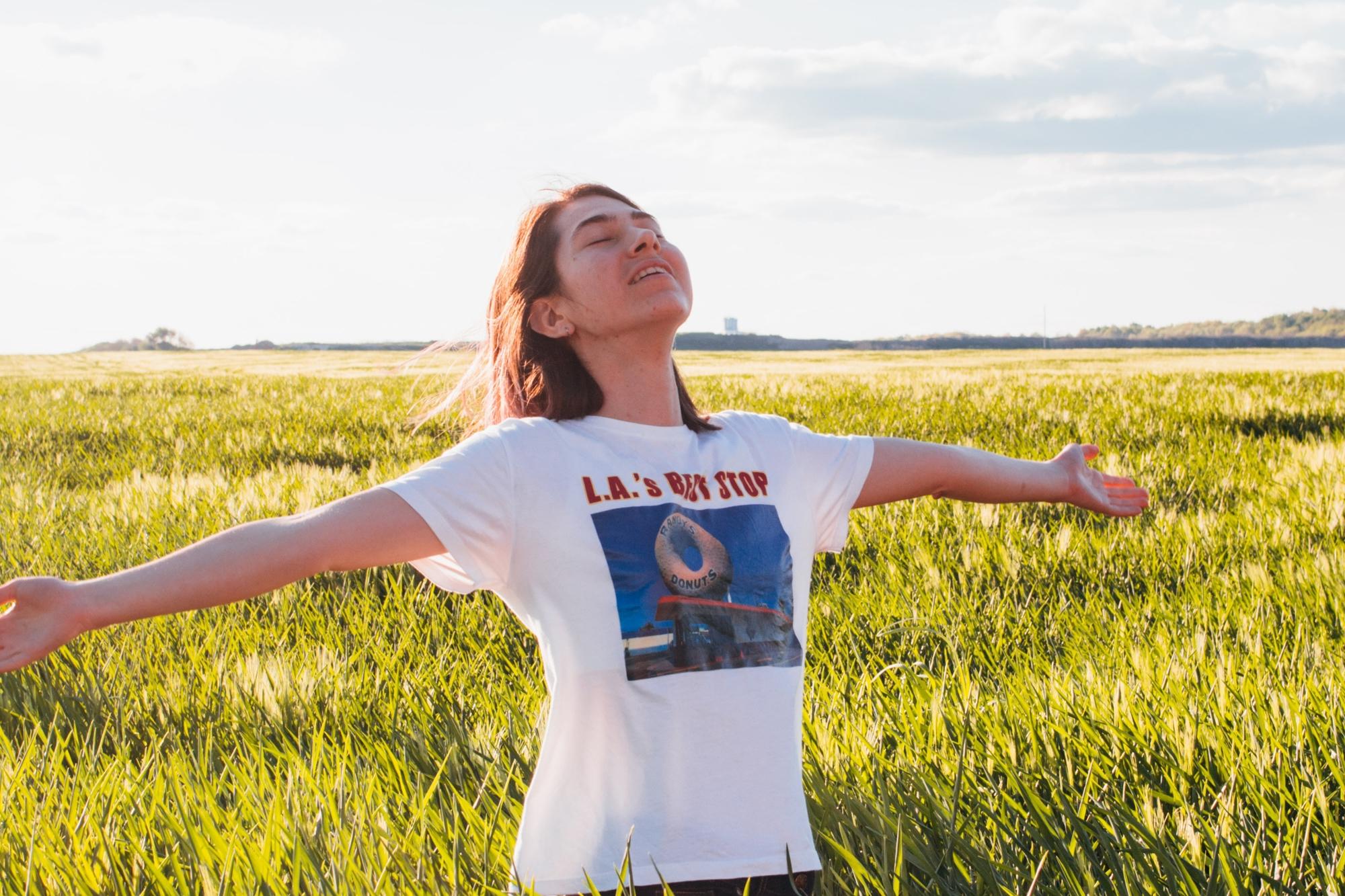En kvinna som står på en äng av högt gräs och sträcker ut armarna i luften och huvudet upp mot himlen.