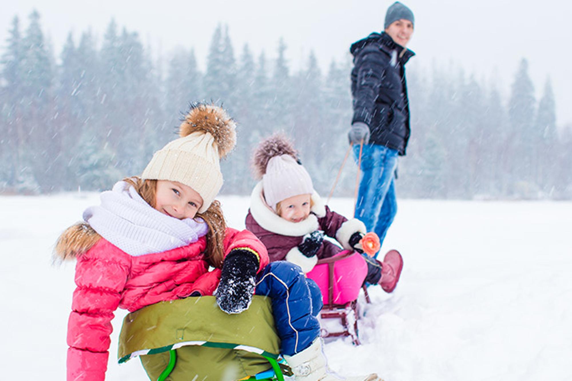 Två glada flickor som sitter i varsin pulka i snön, och deras pappa som drar pulkorna.