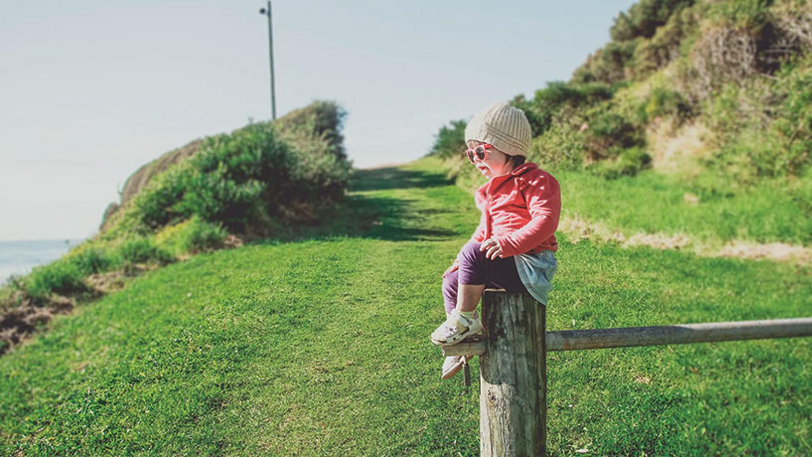 En liten flicka iklädd ytterkläder och ett par solglasögon. Hon sitter på ett staket ute i naturen och tittar framåt, till vänster om kameran.