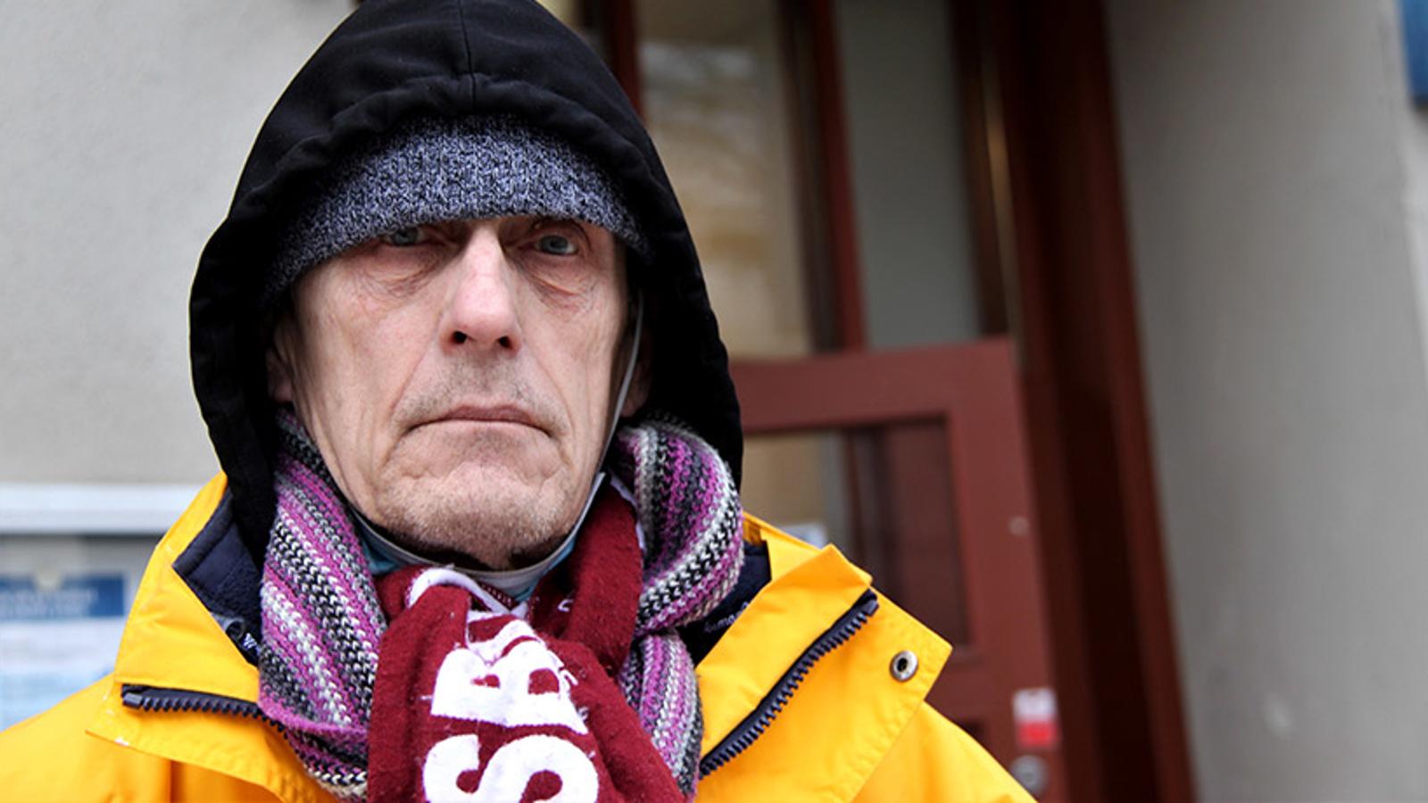 En äldre man, Per-Olof, som står utanför Sociala centret och tittar in i kameran. Han är iklädd en gul jacka, en svart luvtröja, två halsdukar och har en grå mössa på huvudet,