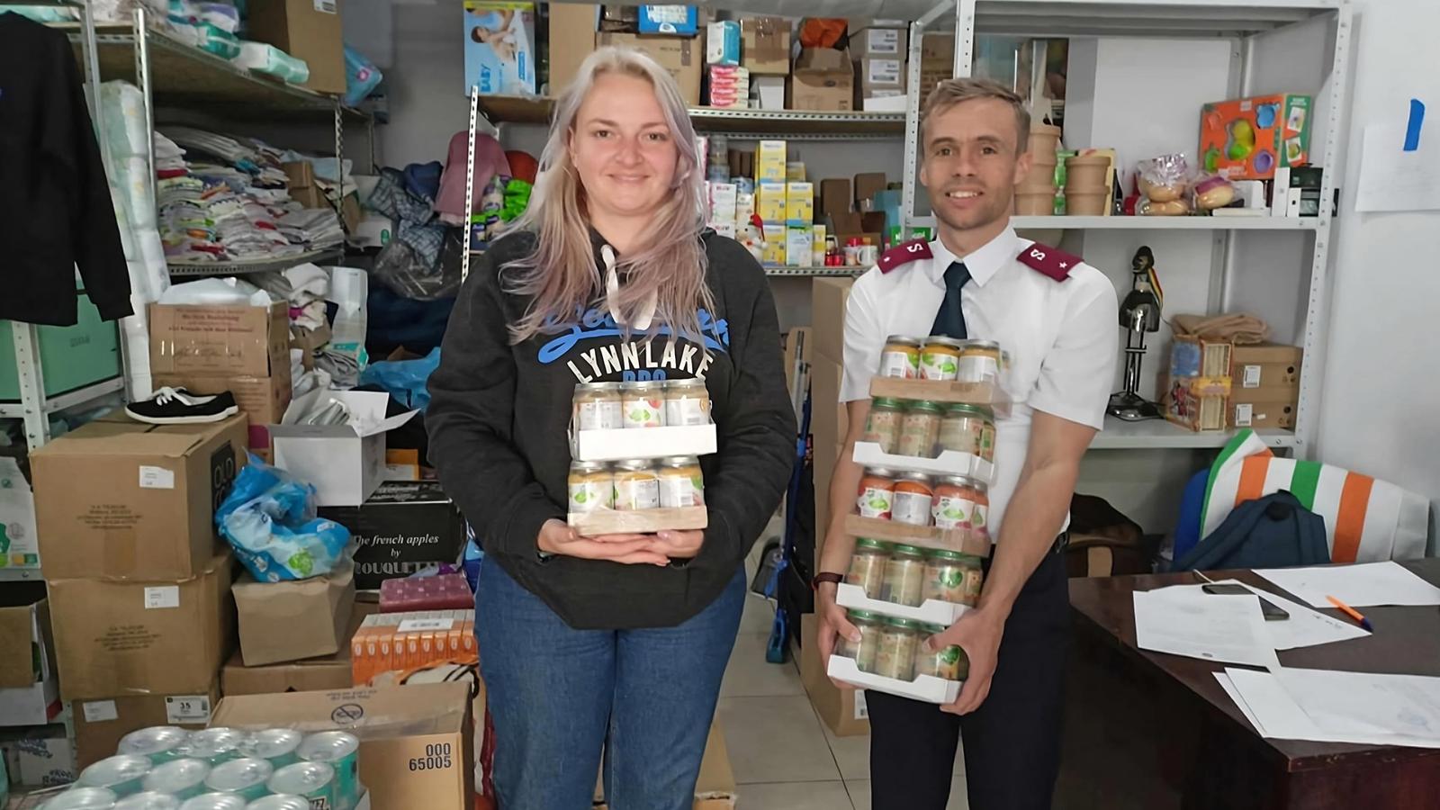 En kvinna och en man står och håller i matförpackningar - i bakgrunden syns ett förråd fullt med matvaror och kläder. Mannen är klädd i Frälsningsarméns uniform.
