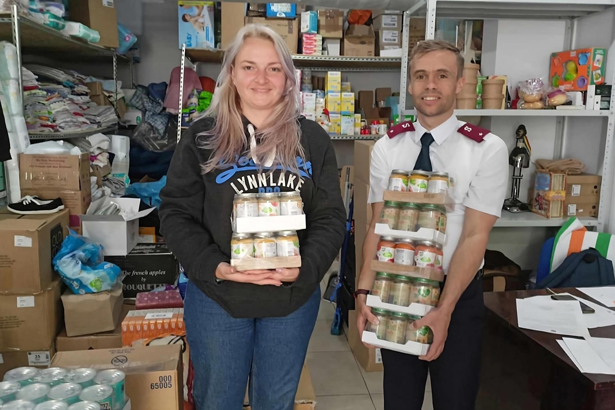 En kvinna och en man står och håller i matförpackningar - i bakgrunden syns ett förråd fullt med matvaror och kläder. Mannen är klädd i Frälsningsarméns uniform.