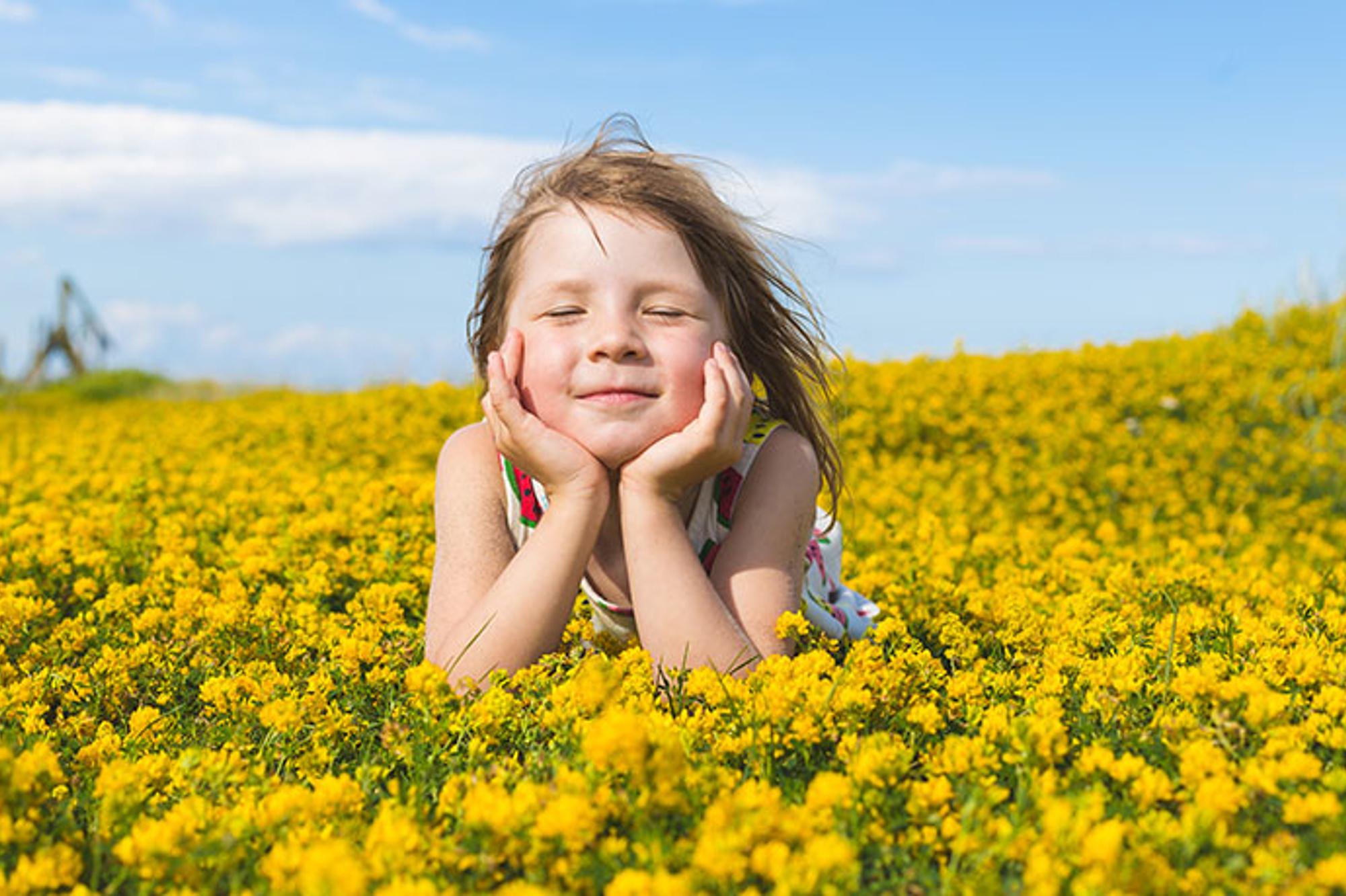 En flicka som ligger på en äng av gula blommor. Hon blundar mot kameran och bakom henne är himlen blå.